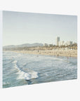 The Offley Green Santa Monica Canvas Wall Art, 'Santa Monica Seaside' showcases a serene beach scene with gentle waves rolling onto the sandy shore. The background features buildings, possibly part of a coastal city like Santa Monica Pier, and is lined with distant mountains under a clear sky. Perfect as beach-themed wall art.