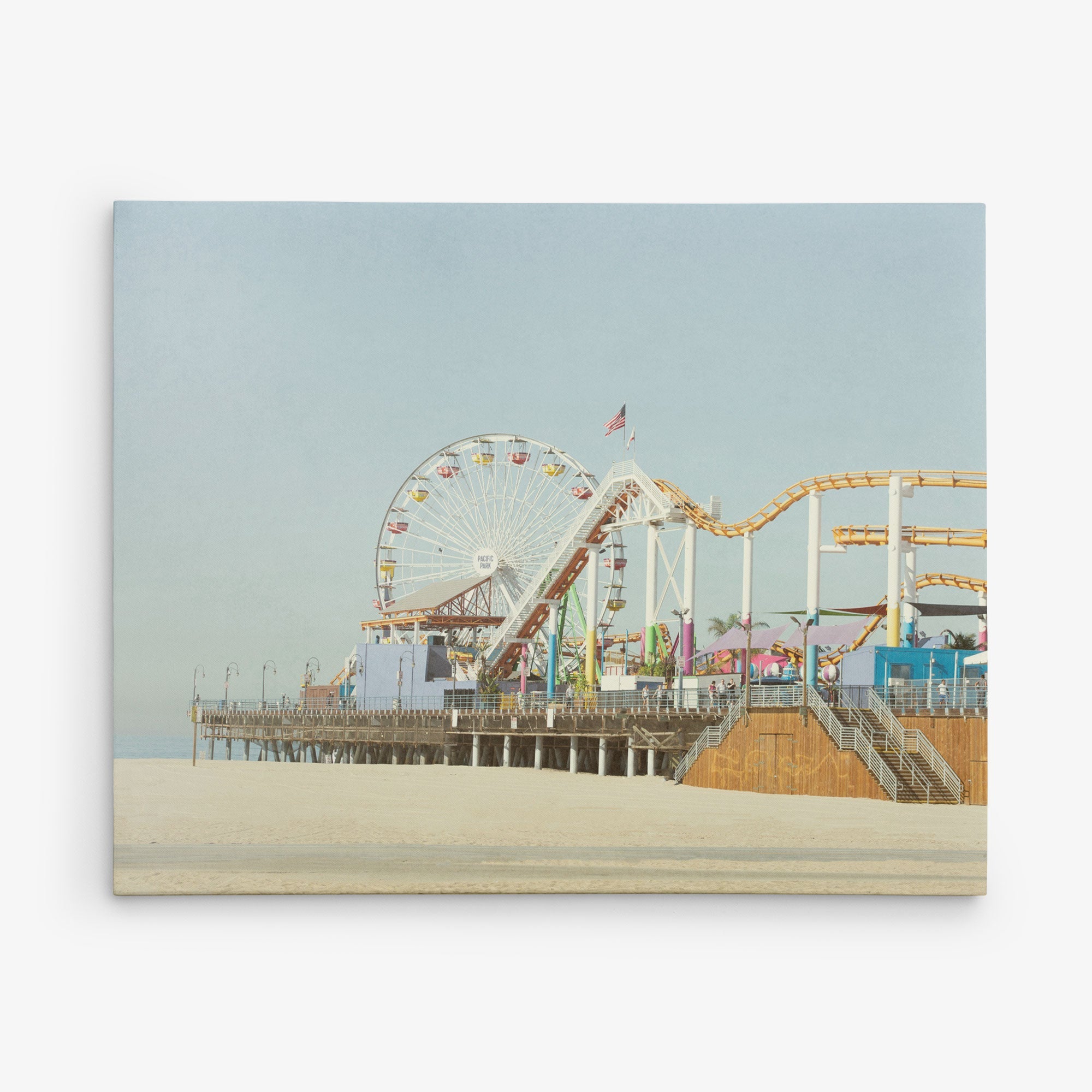 A beach scene featuring a seaside amusement park with a large Ferris wheel, roller coaster, and various other rides and attractions on a pier extending over the sandy beach. The sky is clear and blue, making it perfect for Offley Green's 16x20 California Canvas Print (Choose from 10+ Designs).