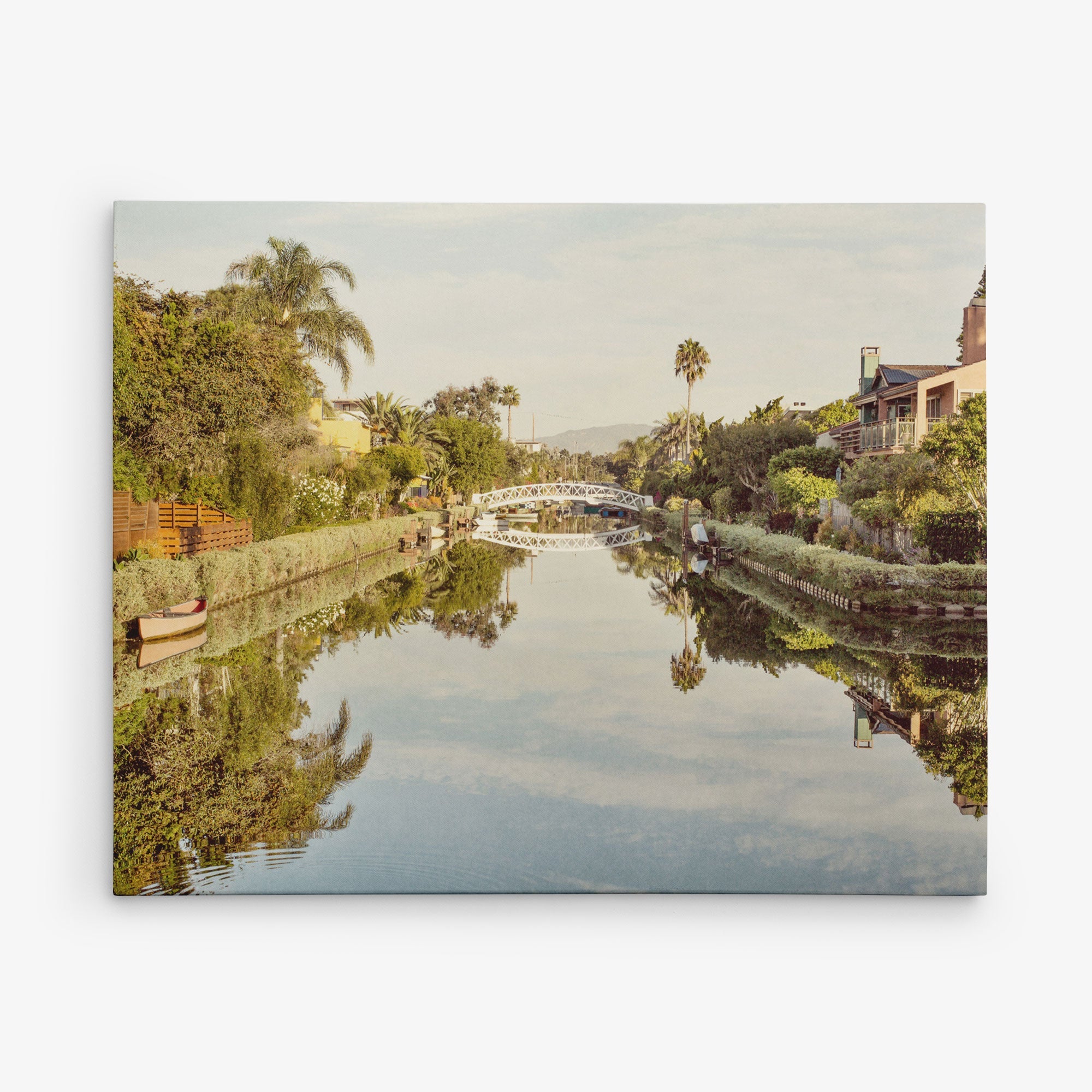 A peaceful canal lined with green trees and shrubs, with a white arched pedestrian bridge in the distance, is the perfect subject for wall art. Houses with gardens border the water, and a few small boats are visible. The clear sky and reflections in the water create a symmetrical scene ideal for Offley Green's 16x20 California Canvas Print (Choose from 10+ Designs).