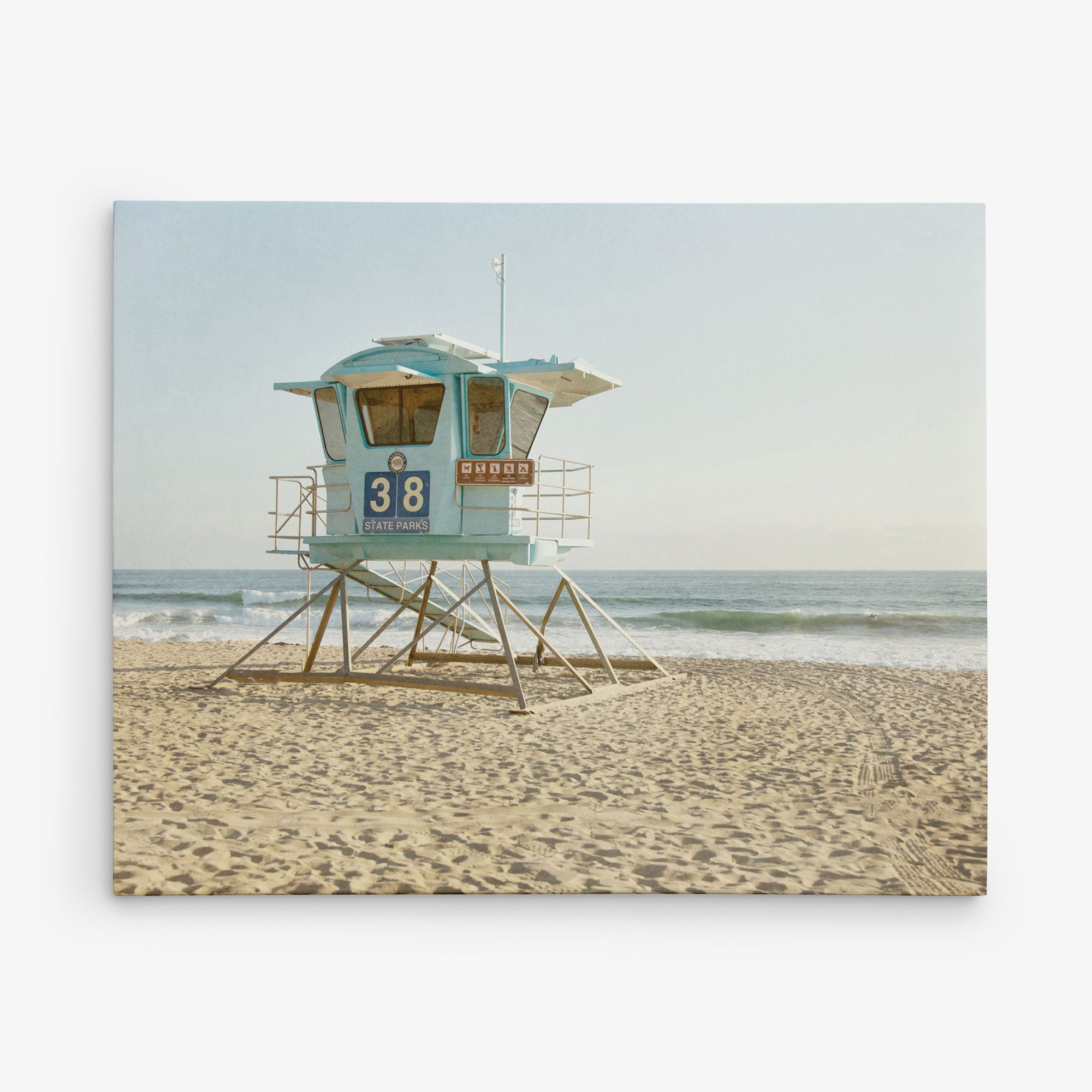 A blue lifeguard tower labeled "38" stands on a sandy beach with the ocean waves gently rolling in the background. The sky is clear and the sun is shining, casting a soft, warm light on this perfect California scene, ideal for an Offley Green 16x20 California Canvas Print (Choose from 10+ Designs).