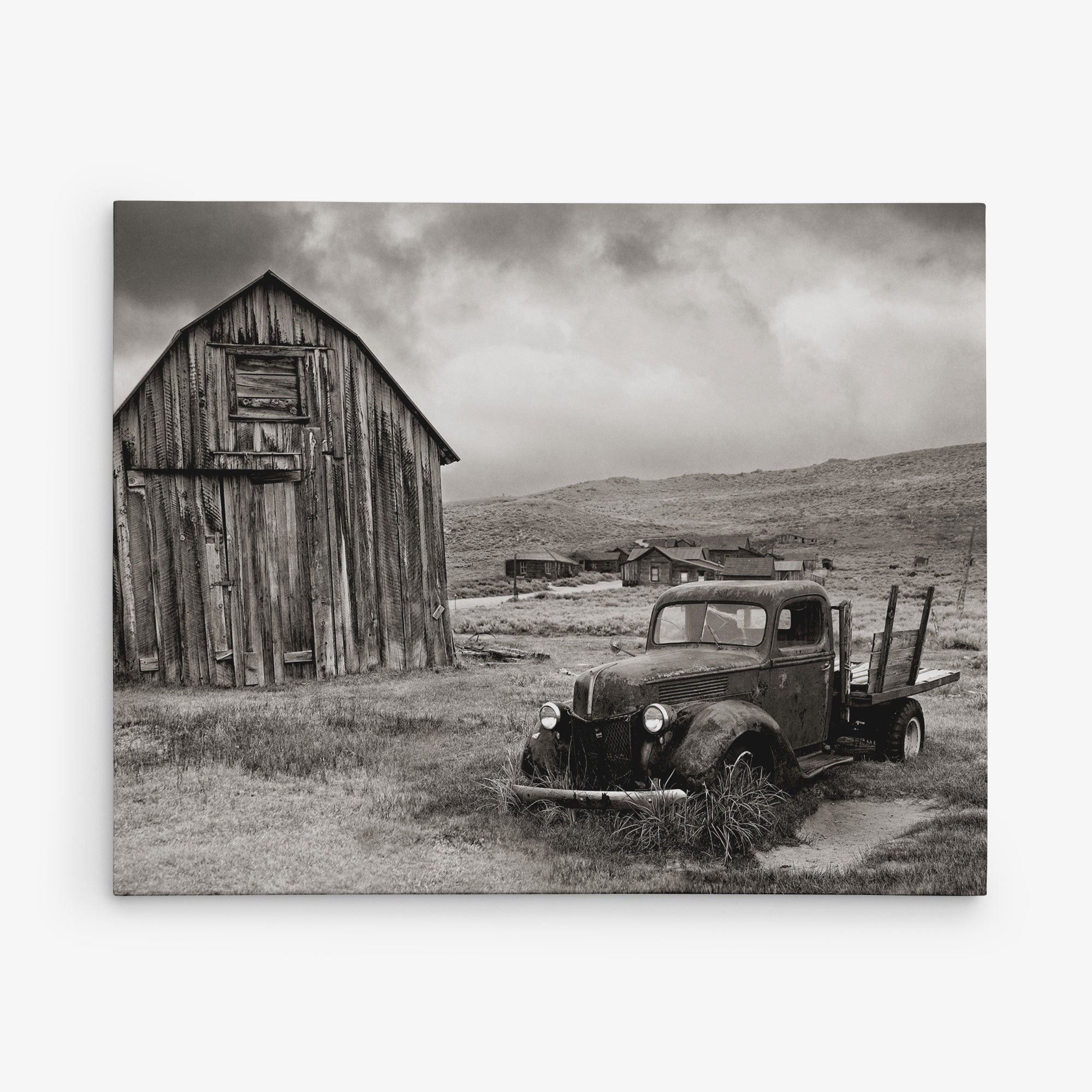A sepia-toned photograph depicts an old, dilapidated truck partially covered in grass, parked in front of a weathered wooden barn. In the background, rustic buildings dot the cloudy sky. This scene of an abandoned rural area makes for stunning wall art or canvas gallery wrap, perfect for those who appreciate Offley Green's 11x14 Rustic Canvas Print (Choose from 10+ Designs).
