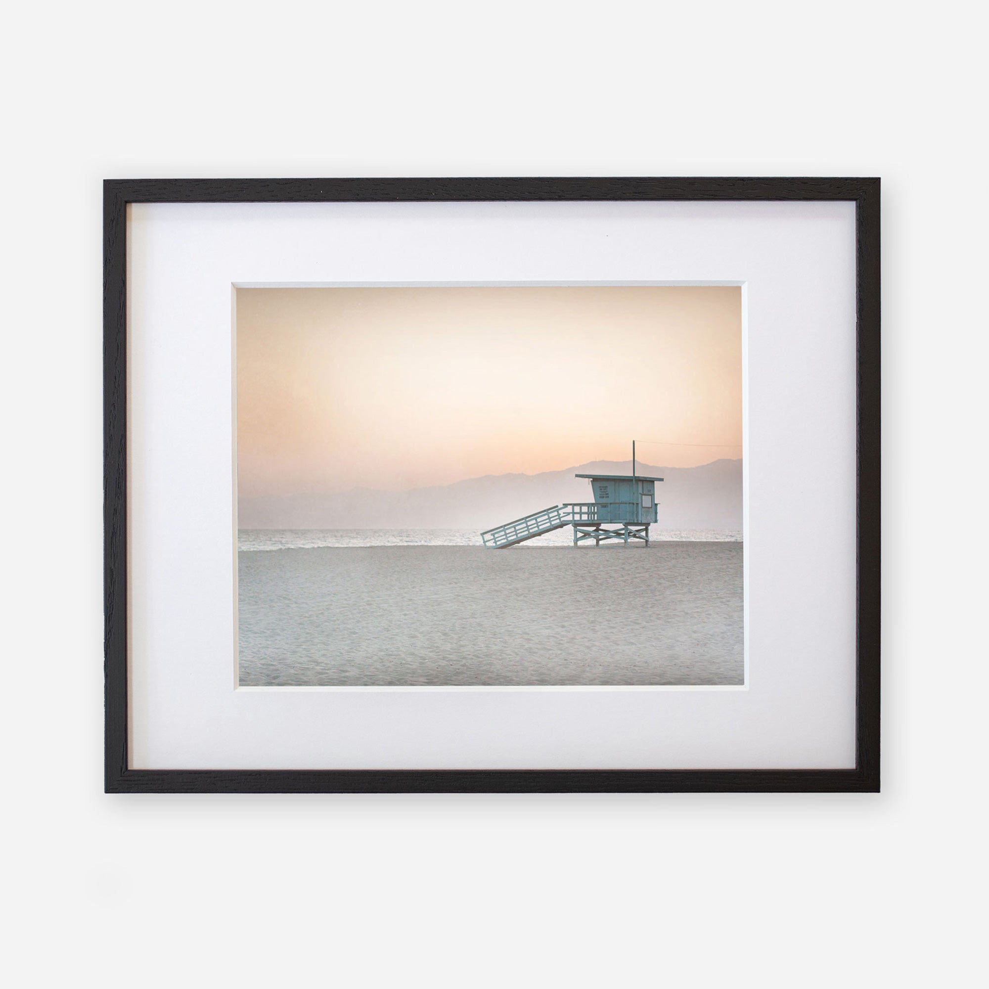 A framed photograph on white archival photographic paper depicting a Pink Coastal Print, 'Lifeguard Tower' by Offley Green on a tranquil beach at sunset, with a soft gradient sky and gentle sea.