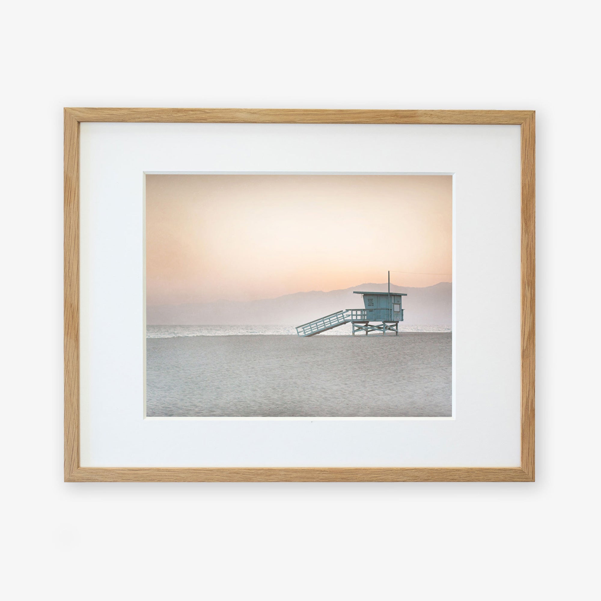 A framed photograph of a Pink Coastal Print, 'Lifeguard Tower' by Offley Green, featuring a serene beach scene at sunrise with a solitary lifeguard station and misty mountains in the background, displayed on a white wall with a non-glossy lustre finish.