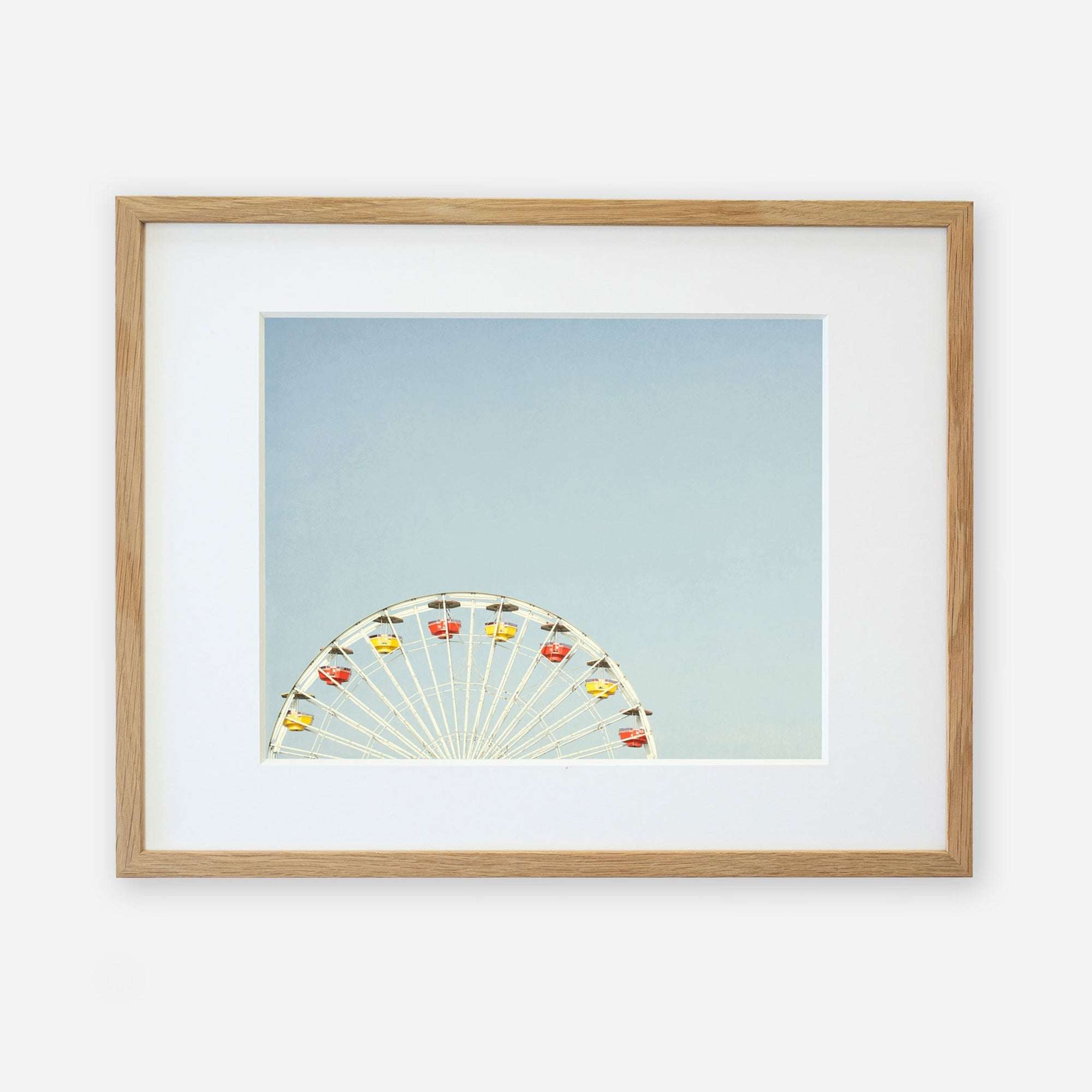 A framed photograph of Ferris Blue, the Santa Monica Pier ferris wheel, partially visible at the bottom, against a light blue sky, displayed in a simple wooden frame with a white mat by Offley Green.