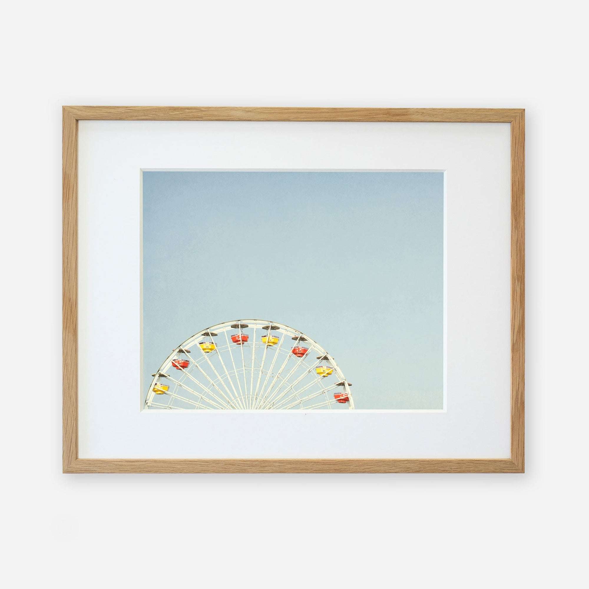 A framed photograph of Ferris Blue, the Santa Monica Pier ferris wheel, partially visible at the bottom, against a light blue sky, displayed in a simple wooden frame with a white mat by Offley Green.