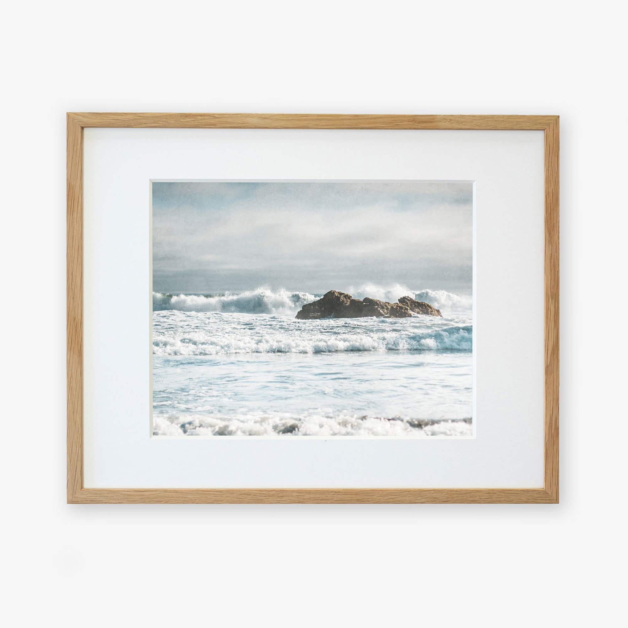 A framed Seascape Coastal Art, 'Surf and Rocks' from Offley Green featuring a serene ocean scene from Pffiefer Beach. In the image, waves crash gently against rocky outcrops under a partially cloudy sky, creating a moody seascape. The frame is wooden with a simple white mat surrounding the picture, printed on archival photographic paper for longevity.