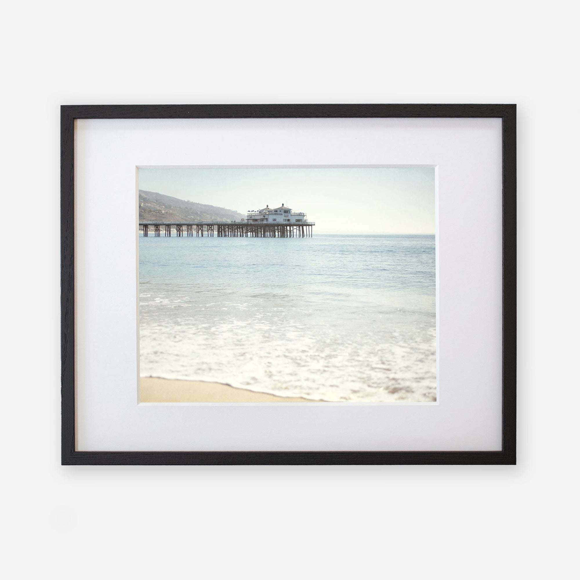 A framed photograph of a serene beach scene, showing gentle waves lapping against the shore with 'Malibu Pier' extending into a calm sea under a clear sky by Offley Green.