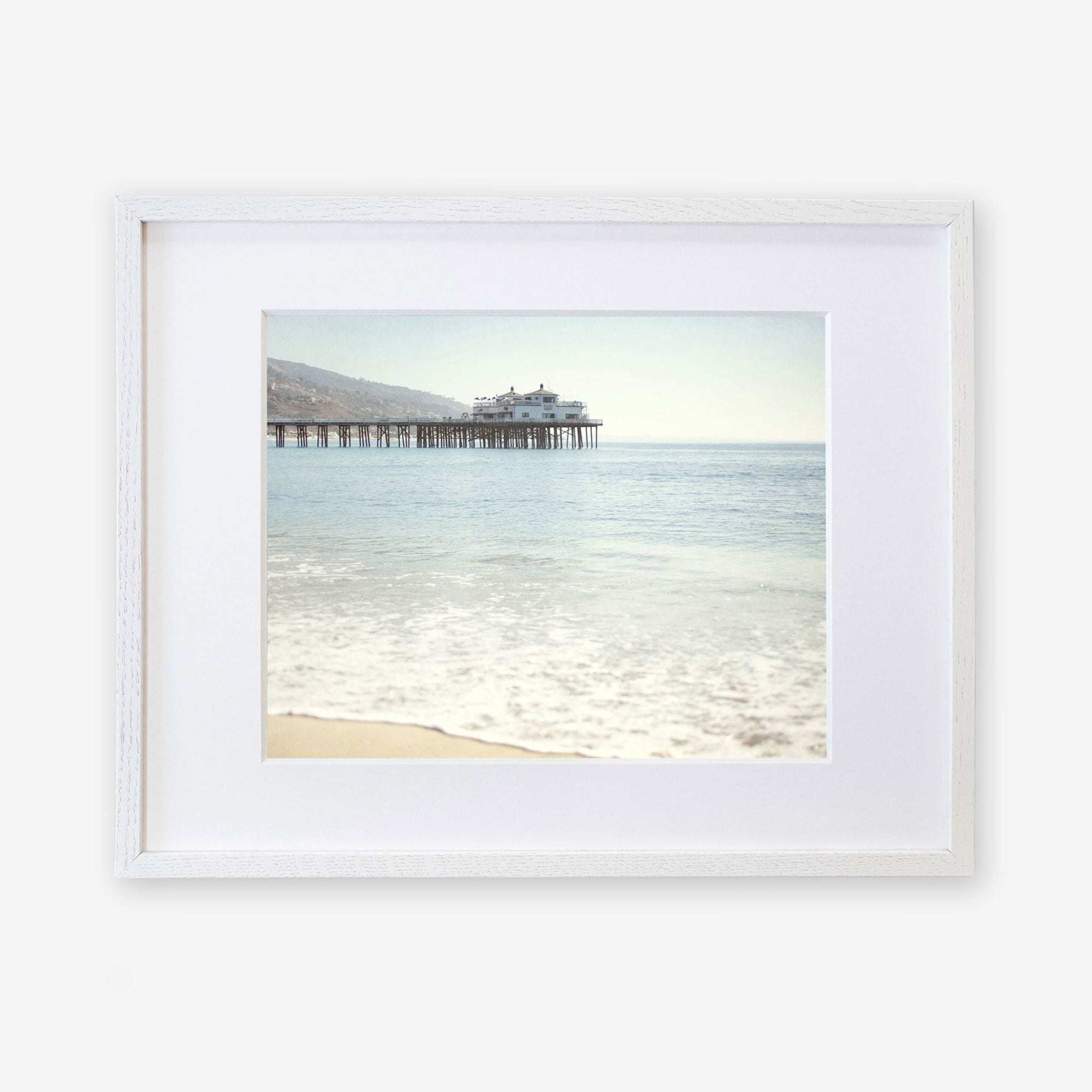 Framed photograph of a peaceful beach with gentle waves lapping at the shore, showcasing 'Malibu Pier' extending into the calm blue ocean under a clear sky - Offley Green's California Beach Print.