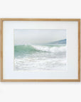 A framed photograph of a wave captured at the moment it breaks on a Southern California beach, with a foamy crest and misty backdrop, hung on a plain white wall. Offley Green's Coastal Print of a Breaking Wave 'Breaking Surf'