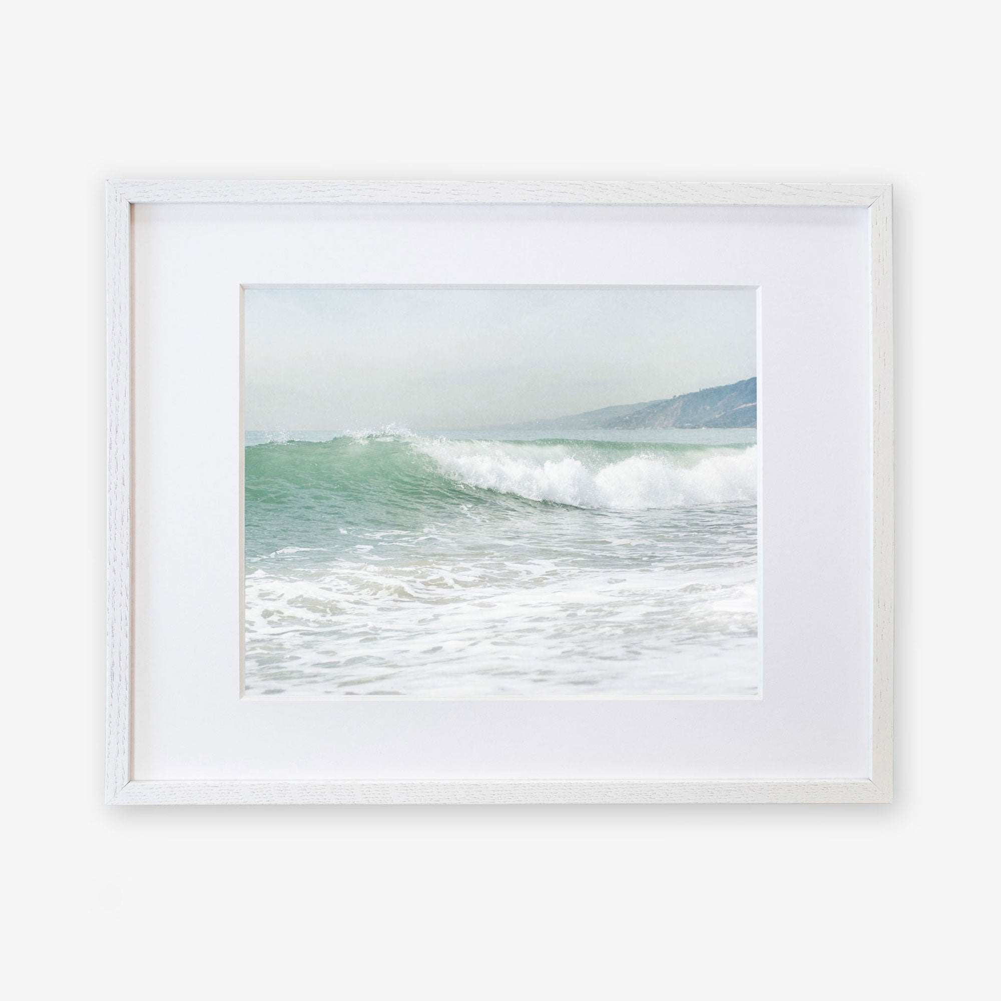 A framed Breaking Surf print of a vibrant ocean wave cresting at a Southern California beach, displayed in a simple white frame against a white background by Offley Green.