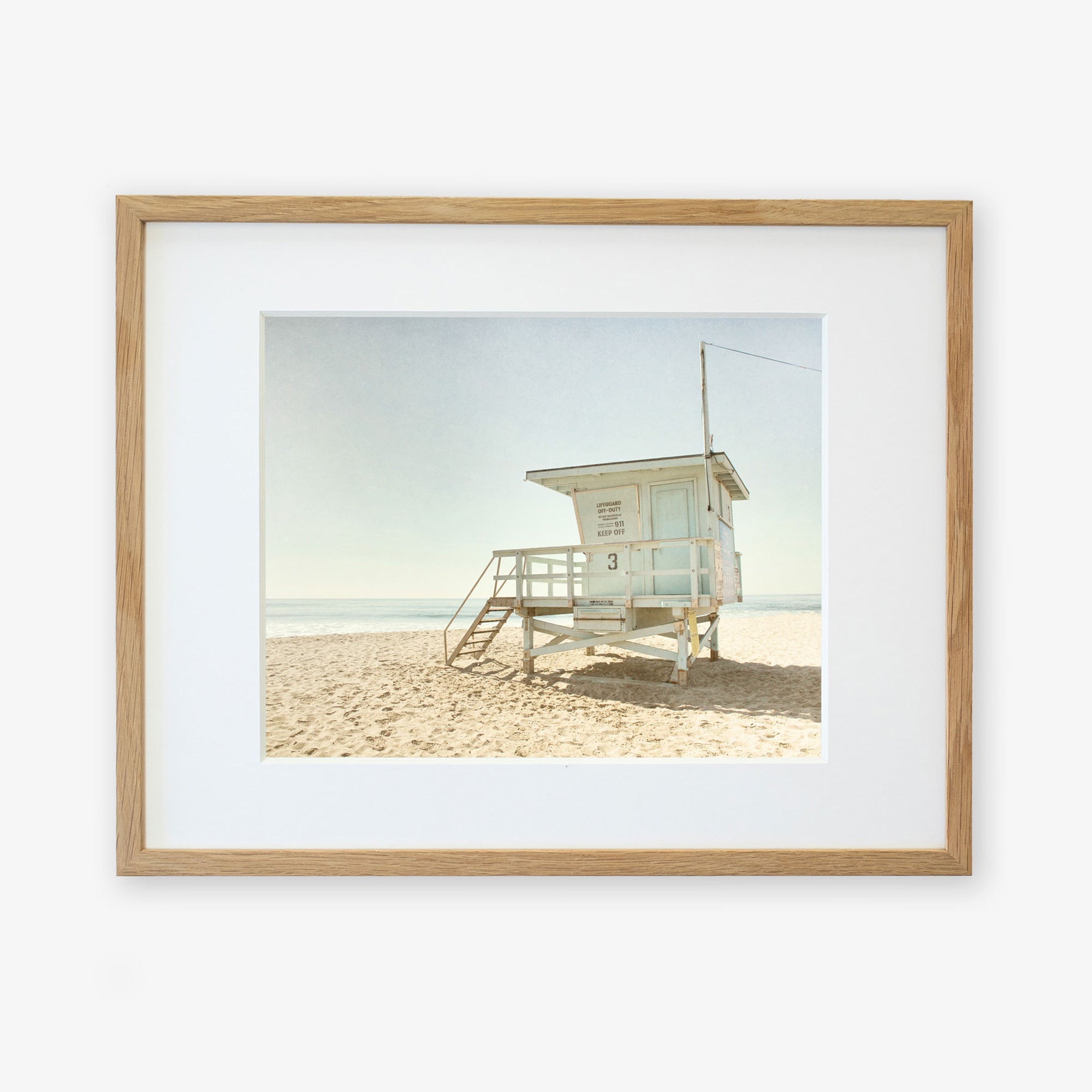 A framed photograph depicting a solitary beach lifeguard station on sandy shores under a clear sky. The guard station, numbered &quot;3&quot; and located in Malibu, features a small set of steps leading to Offley Green&#39;s California Summer Beach Art, &#39;Malibu Lifeguard Tower&#39;.