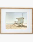 A framed photograph depicting a solitary beach lifeguard station on sandy shores under a clear sky. The guard station, numbered "3" and located in Malibu, features a small set of steps leading to Offley Green's California Summer Beach Art, 'Malibu Lifeguard Tower'.