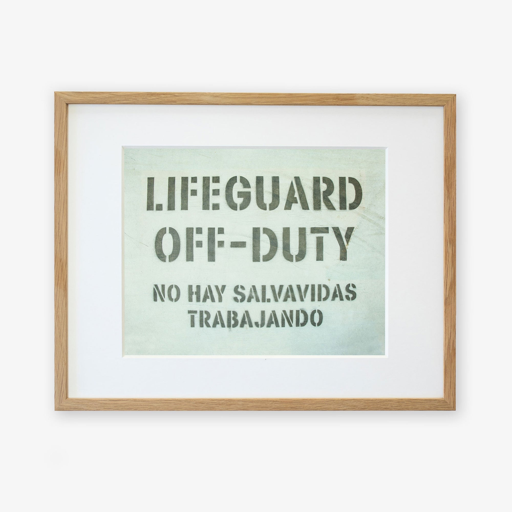 A framed Teal Blue Surfer Beach Print, 'Off Duty' by Offley Green with the text "LIFEGUARD OFF-DUTY" in large letters and "NO HAY SALVAVIDAS TRABAJANDO" in smaller letters beneath, indicating no lifeguards are currently on duty. The text is in black on a light background, printed on archival photographic paper. The frame is light wood.