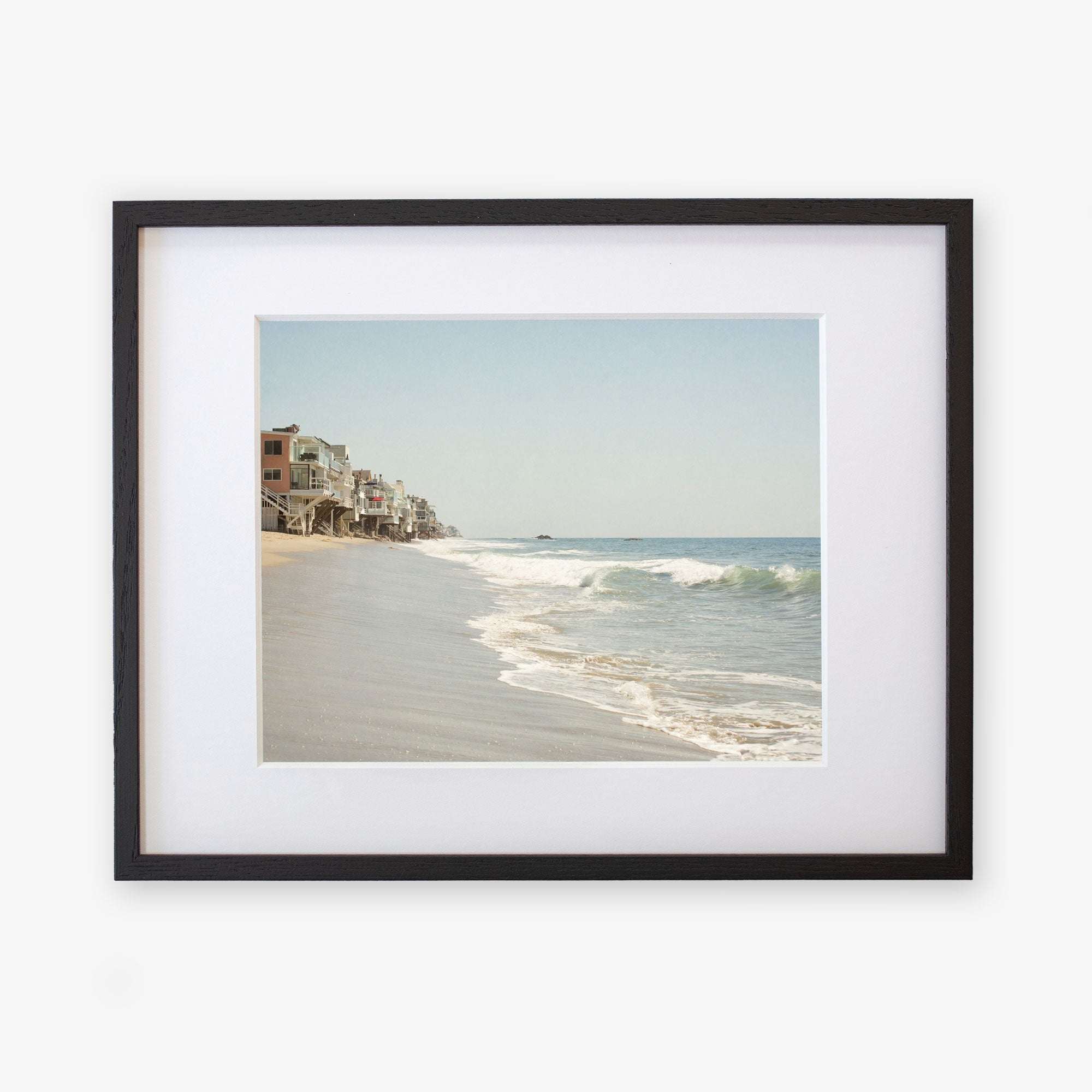 A framed photograph of a peaceful Malibu coastline showing gentle waves lapping at the shore and a row of seaside houses extending into the distance, printed on archival photographic paper by Offley Green.