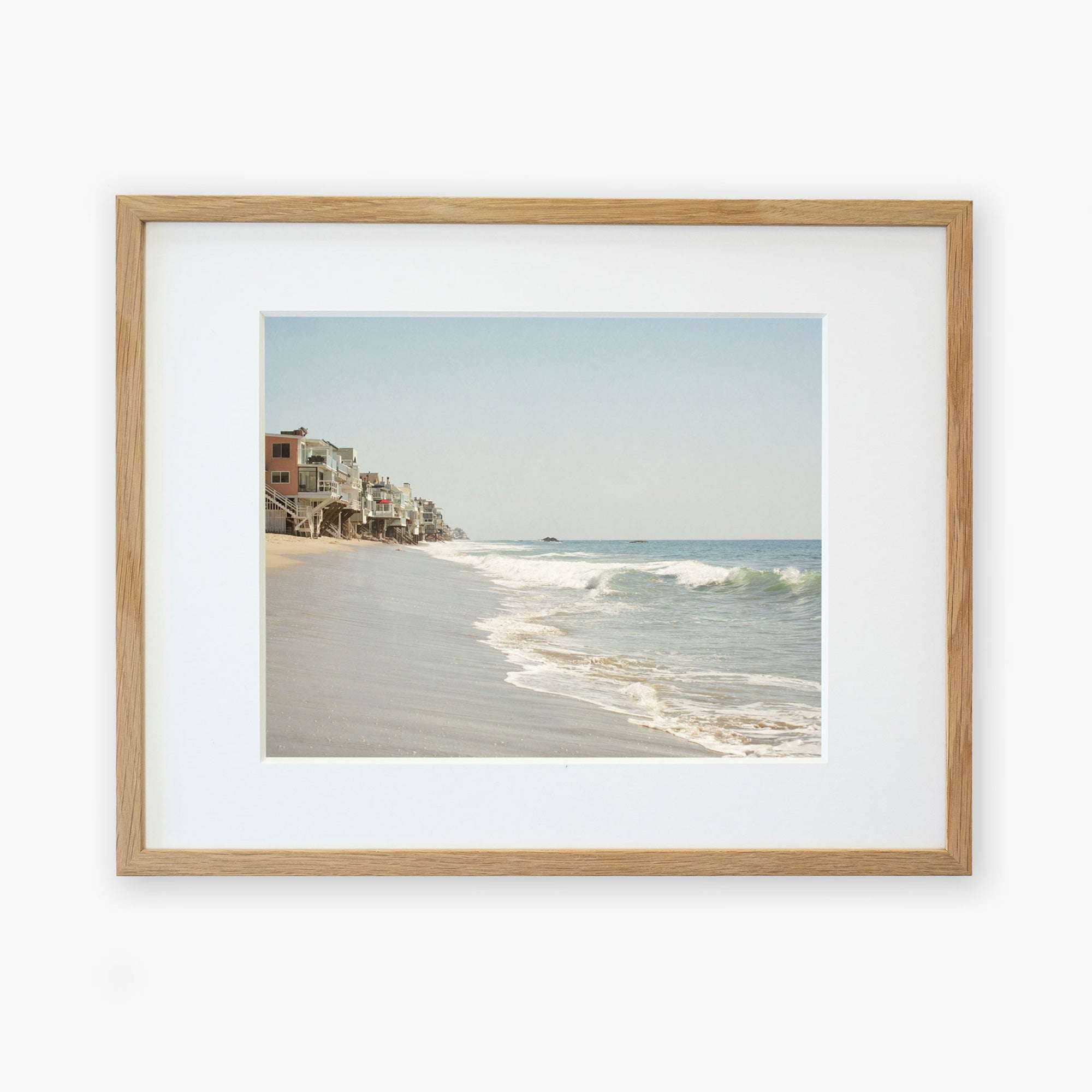 A framed photograph of a Malibu Beach House Print, 'Ocean View' by Offley Green showing gentle waves lapping onto a sandy shore with a line of houses extending along the beachfront.