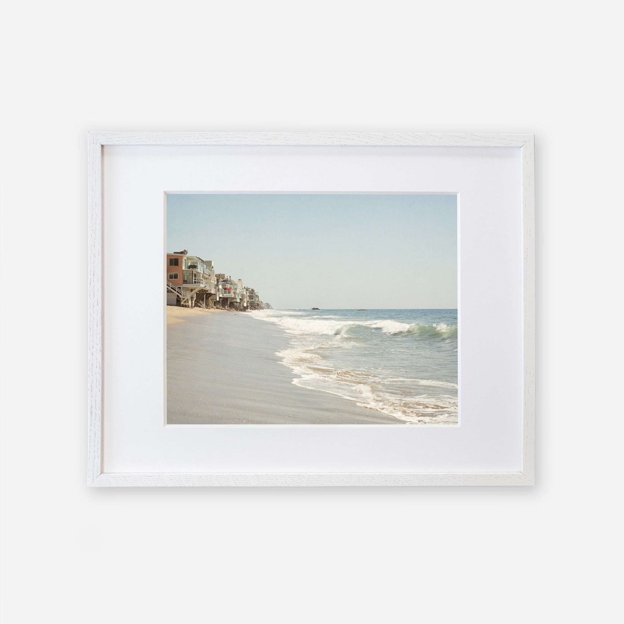 A framed Malibu Beach House Print, 'Ocean View' of a serene Malibu coastline with waves gently lapping at the shore and a row of buildings extending along the coast under a clear sky by Offley Green.