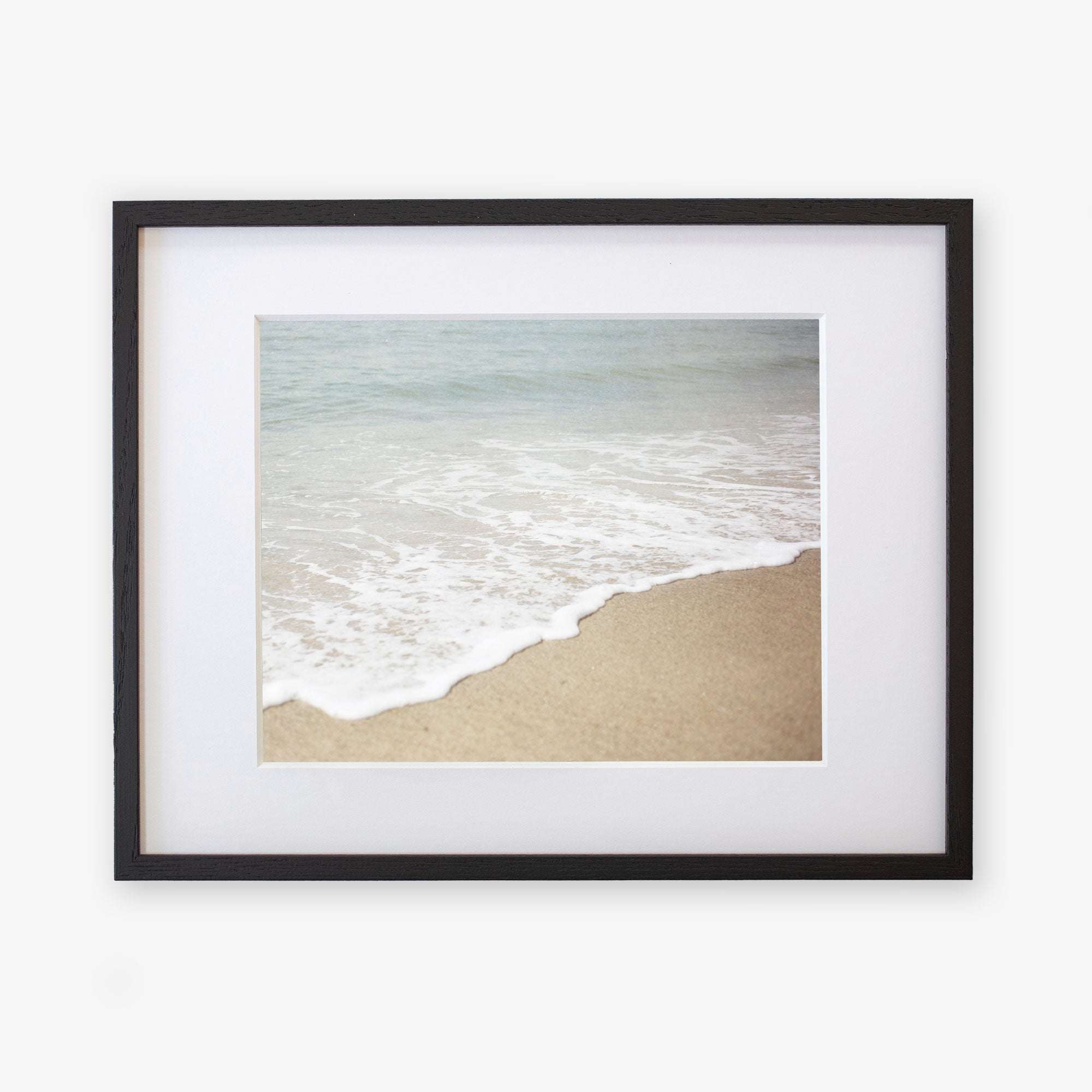 A framed Beach Waves Print, 'Chasing Surf' of a California beach scene, showing gentle waves lapping onto a sandy shore, mounted on archival photographic paper within a black frame by Offley Green.