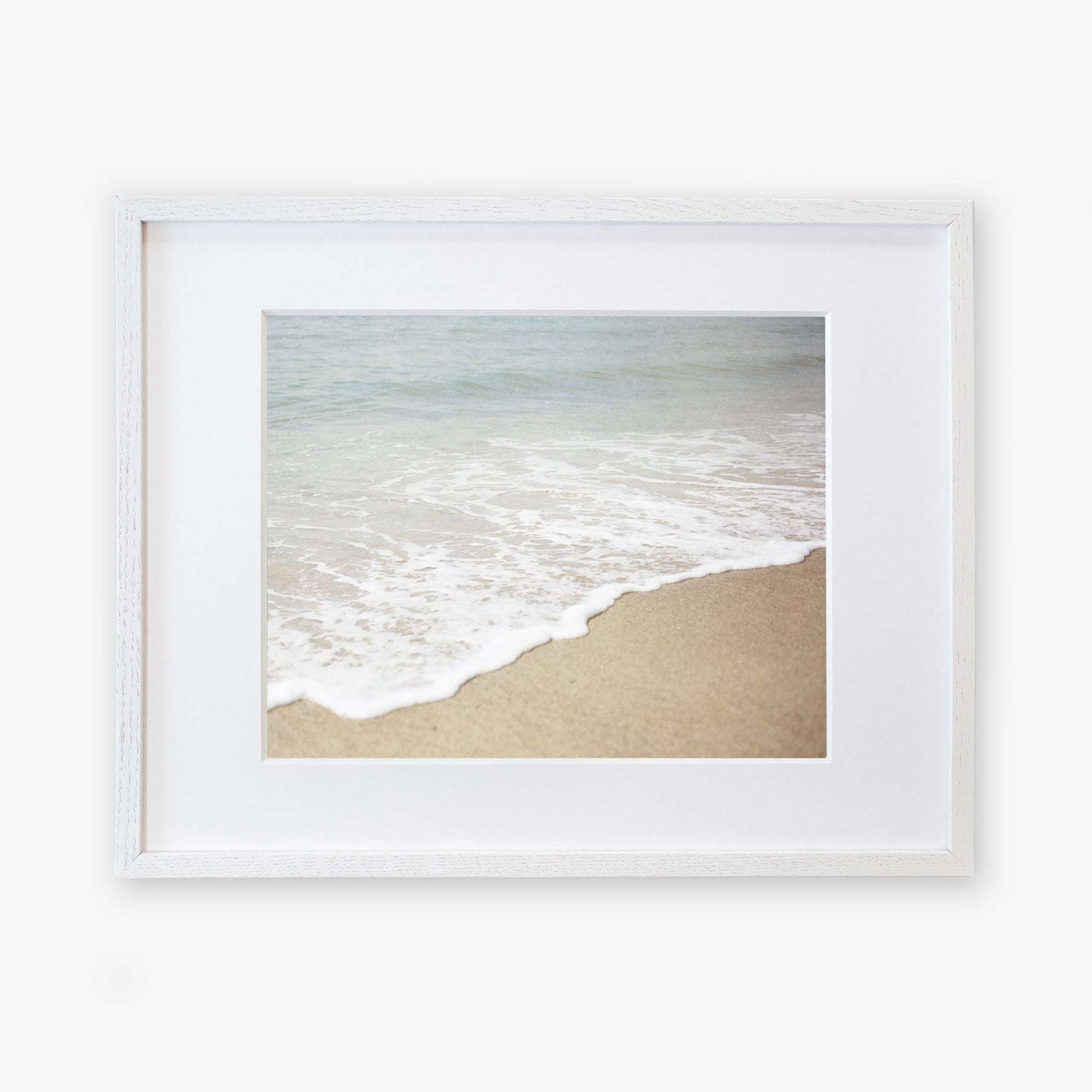 A framed photograph of a California beach with gentle waves lapping at the shore, set against a serene ocean backdrop. The frame is white and mounted on a white background featuring the Offley Green Beach Waves Print, 'Chasing Surf'.