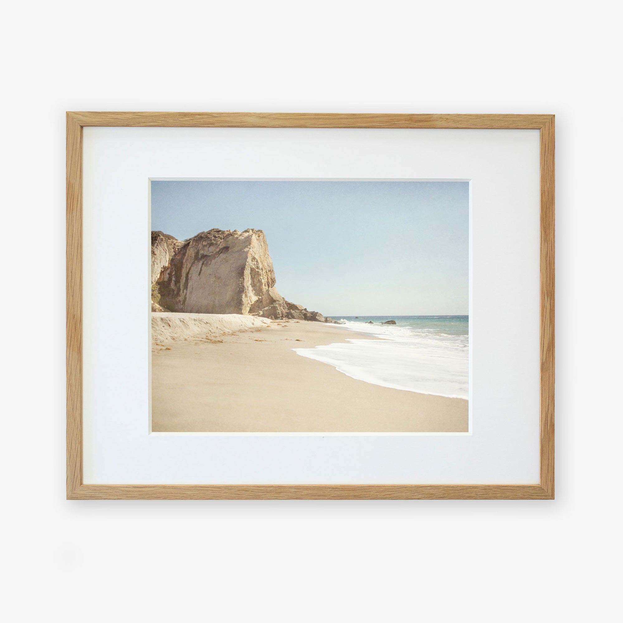 Offley Green's California Malibu Print, 'Point Dume' depicts a serene beachscape featuring a sandy beach with gentle waves at Point Dume, flanked by a large, rugged cliff under a clear sky.