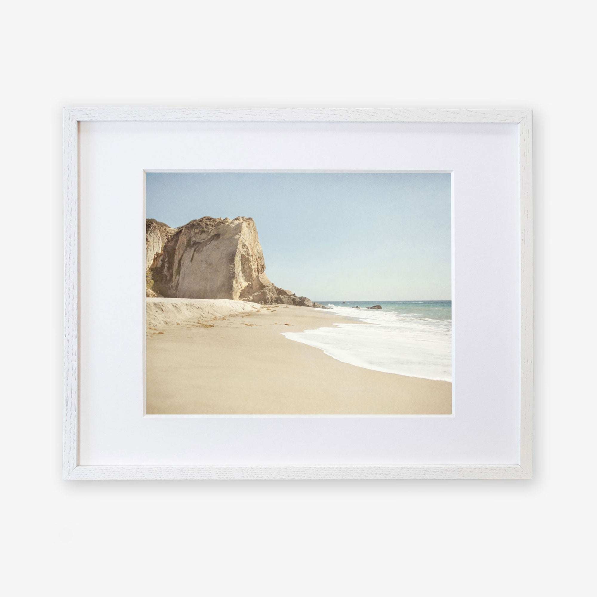 Framed photograph of a serene California Malibu Print, 'Point Dume' beach scene with a prominent rocky cliff on the left and gentle waves washing onto the sandy shore under a clear sky by Offley Green.