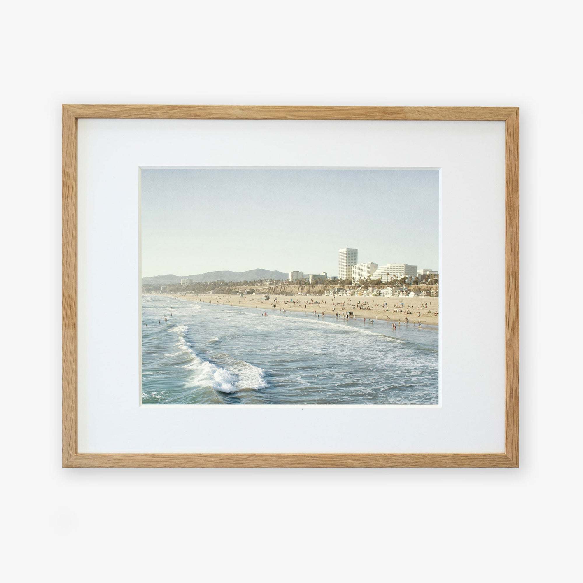Framed photograph of a bustling beach scene with waves crashing along the shoreline at Santa Monica Pier and a crowded sandy beach, set against a backdrop of a city skyline by Offley Green in their Santa Monica Print, 'Santa Monica Seaside'.