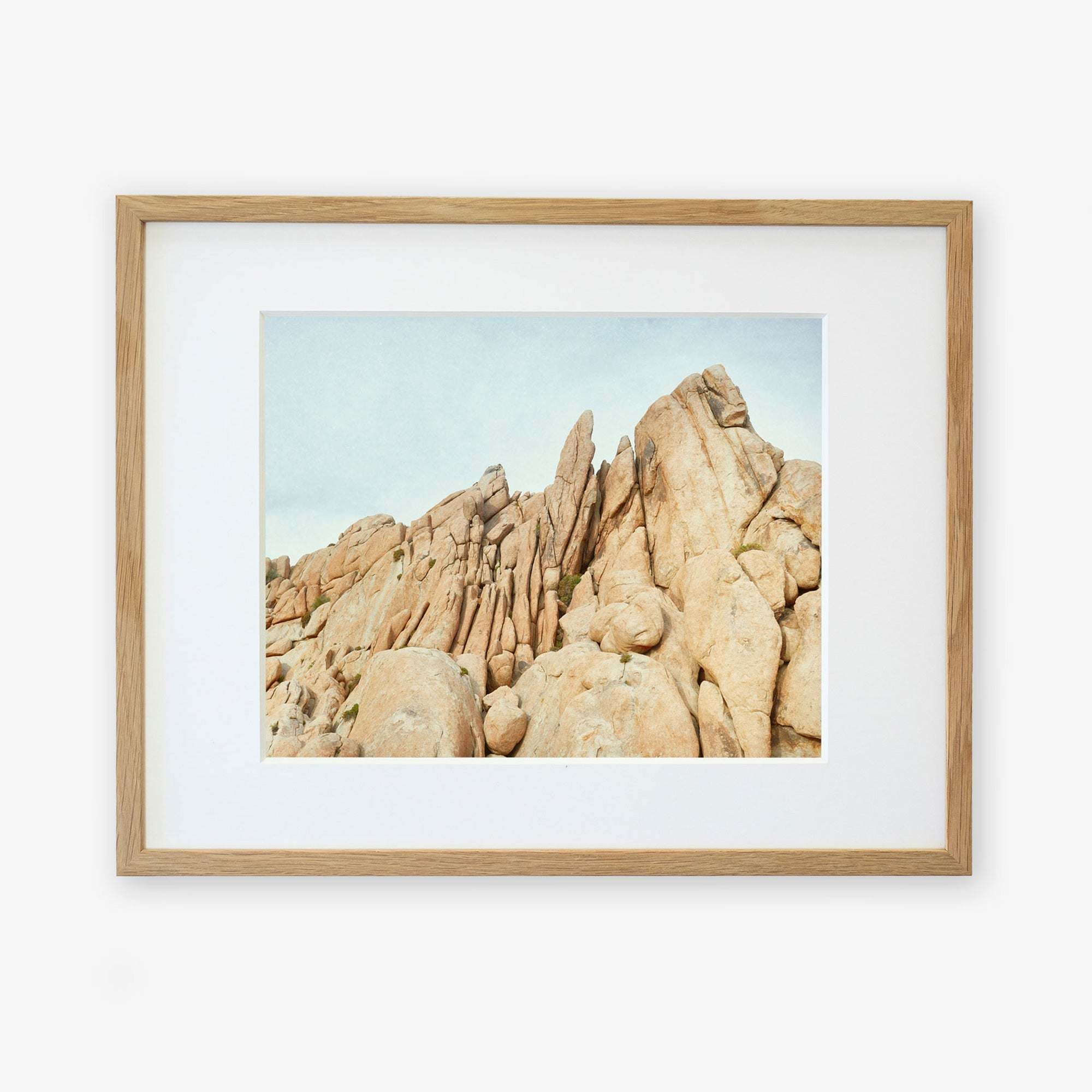 Framed photograph of a Offley Green Joshua Tree Print, 'Joshua Rocks' desert landscape, featuring tall, jagged rock formations under a clear blue sky, displayed in a natural wood frame with a white mat border.