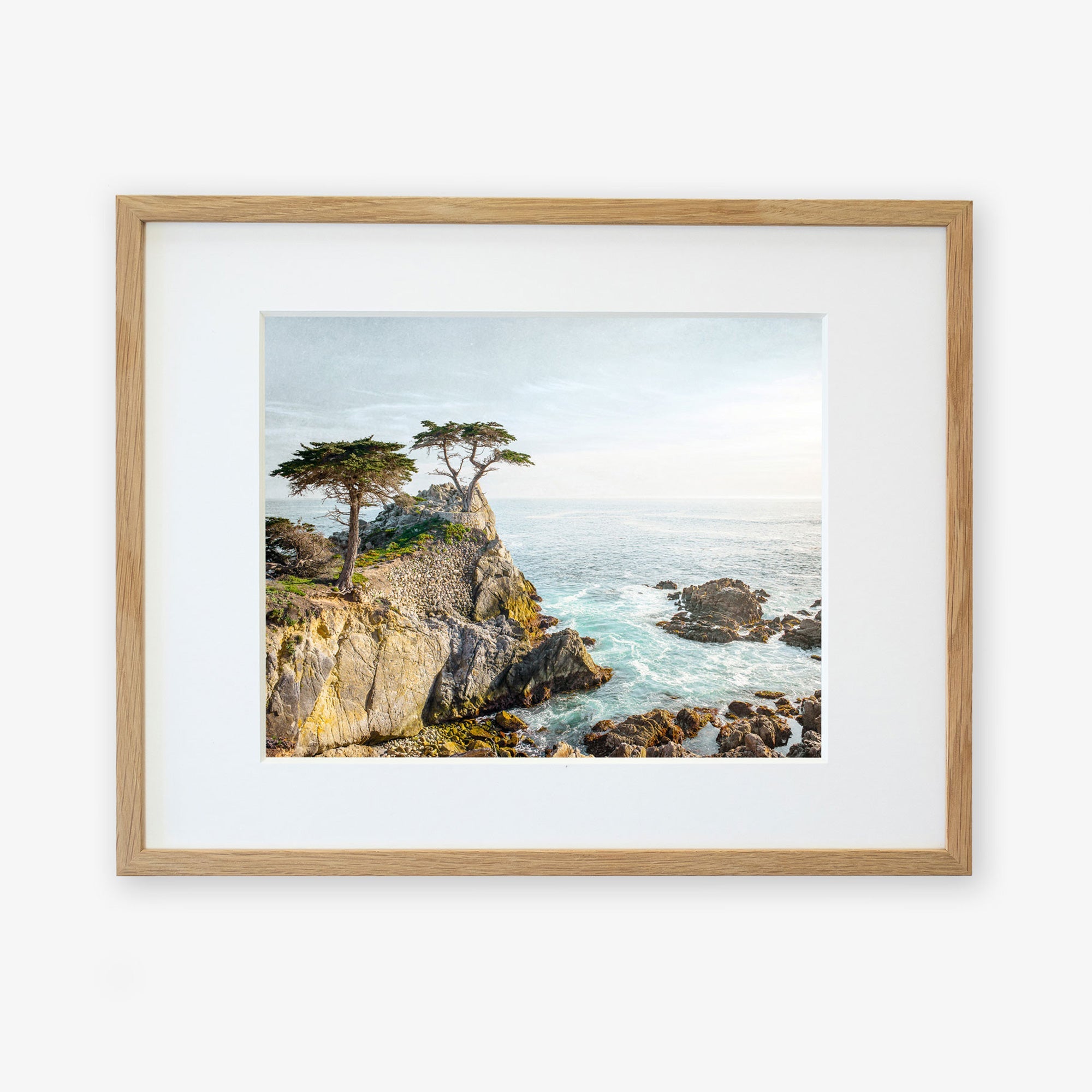 A framed California Coastal Print, &#39;Lone Cypress&#39; by Offley Green, depicting a rugged coastline with several trees atop a cliff overlooking the ocean at Pebble Beach, displayed against a plain white background.