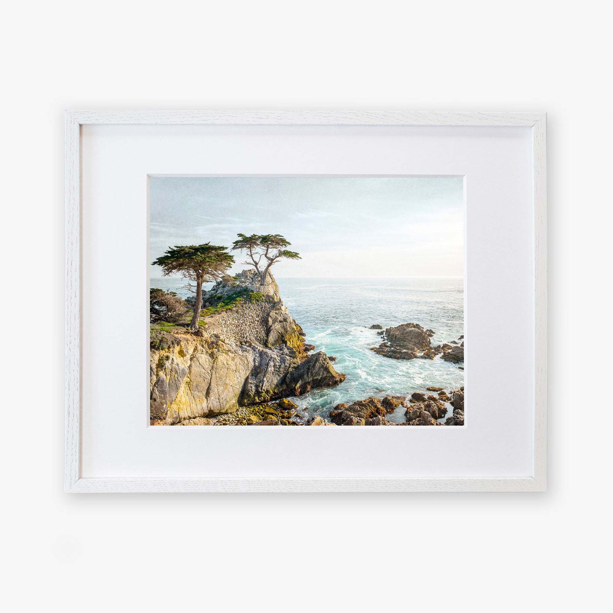 A framed painting of a California Coastal Print, 'Lone Cypress' with rugged cliffs, choppy sea, and windswept trees under a bright sky at Pebble Beach, displayed on a white background by Offley Green.