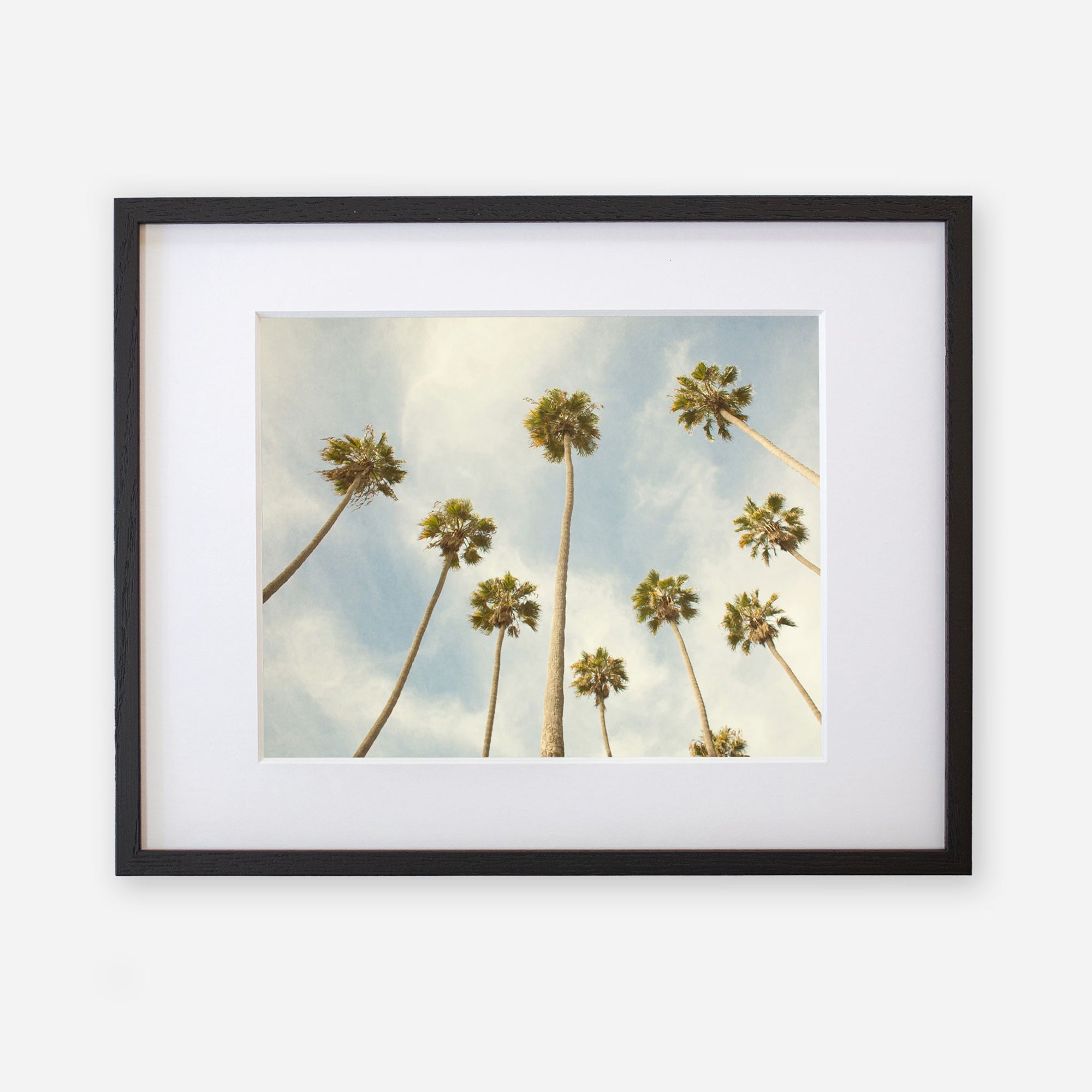 Framed artwork featuring a view of tall California palm trees from below against a cloudy blue sky background, printed on archival photographic paper with a non-glossy lustre finish is the Offley Green Palm Tree Print, California Beach Scene &#39;Reach for the Palms&#39;.