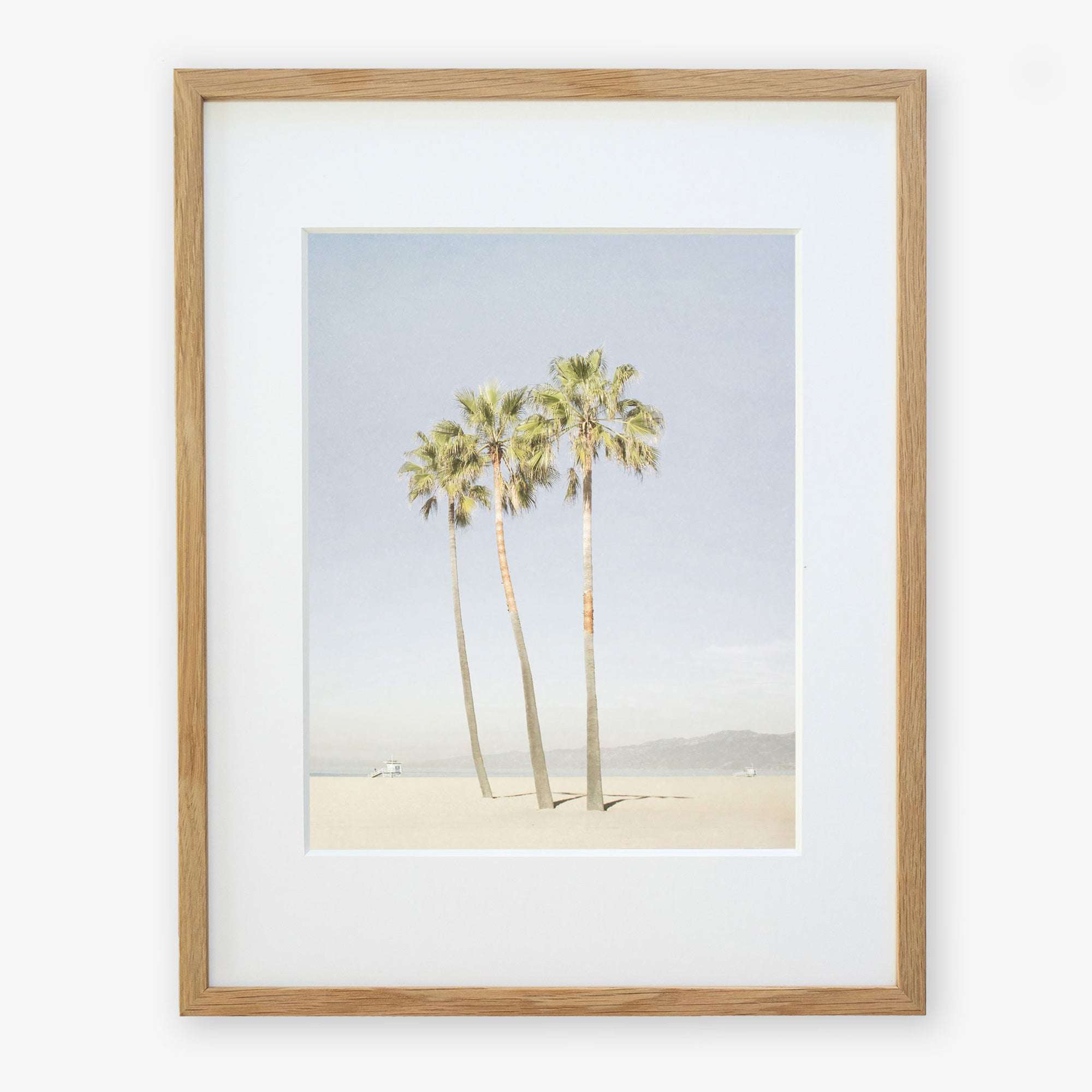 Framed artwork depicting three tall palm trees under a clear sky, with a small figure and a dog at the base, possibly on Venice Beach. The frame is simple and made of light wood. This is the Offley Green California Venice Beach Print, 'Three Palms'.