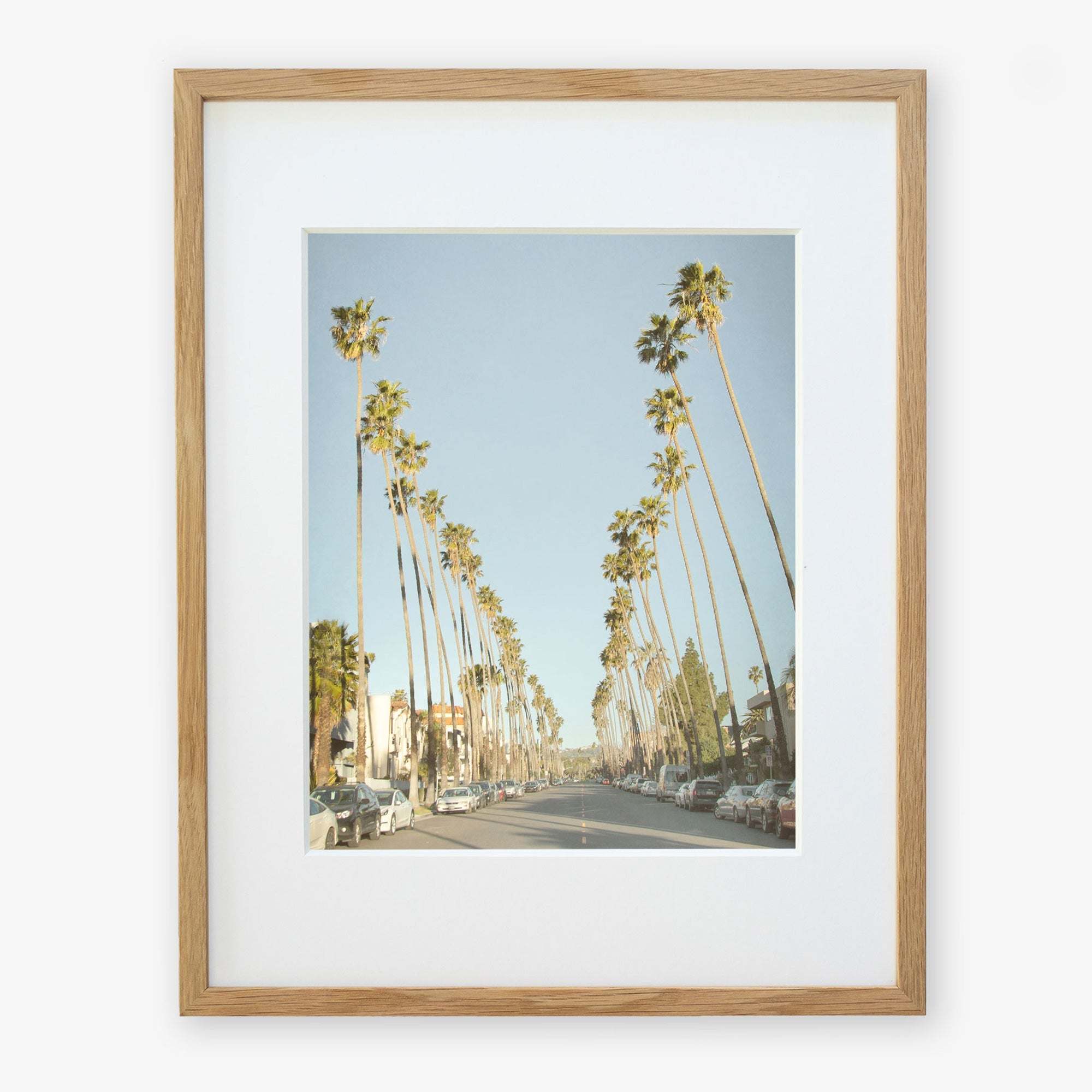 A framed photograph depicting Los Angeles Palm Tree Lined Street 'Sunset Boulevard Dreams', viewed from the perspective of the center of the road, under a clear blue sky by Offley Green.