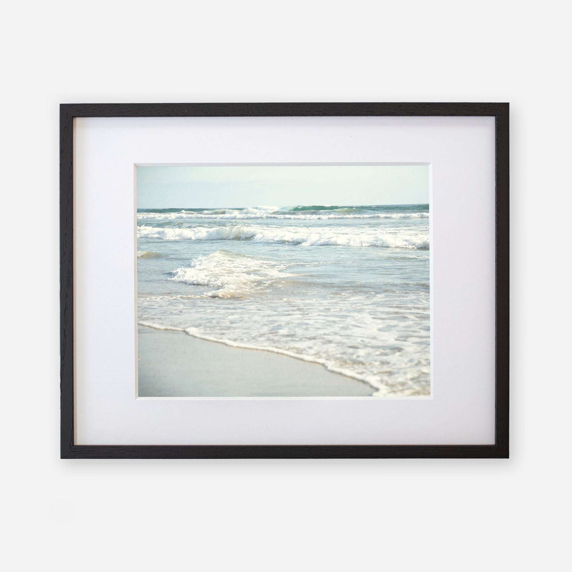 A framed photograph of a serene scene at Carlsbad State Beach, displaying gentle waves breaking near the shore with a clear sky in the background. The frame is black with a white mat featuring the Coastal Beach Print in California 'Surf and Sun' by Offley Green.
