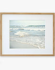 A framed photograph of a tranquil Carlsbad State Beach scene, with gentle waves lapping at the shore under a clear sky. The frame is simple and made of light wood, mounted on a white Offley Green Coastal Beach Print in California 'Surf and Sun'.