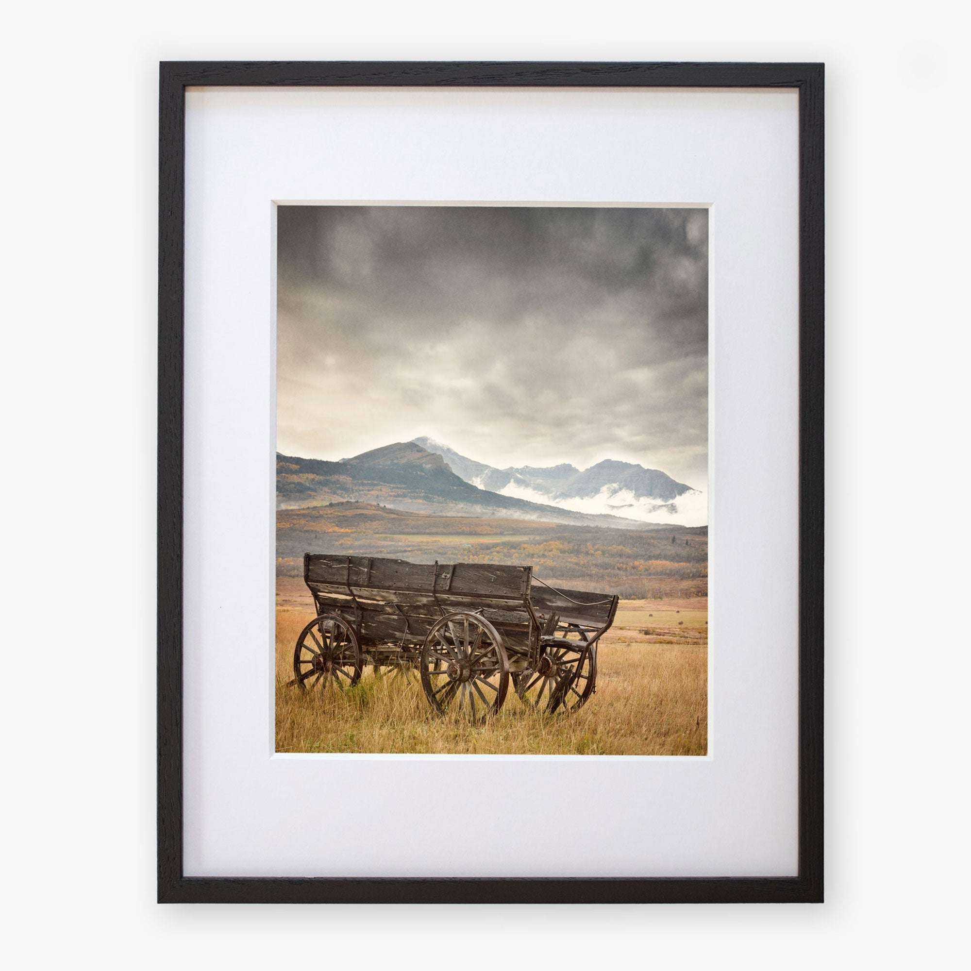 The Offley Green 'Old Wagon' Rustic Farmhouse Wall Art captures a serene scene of a weathered wagon in an open field, accentuated by snow-capped mountains and a cloudy sky for timeless charm.