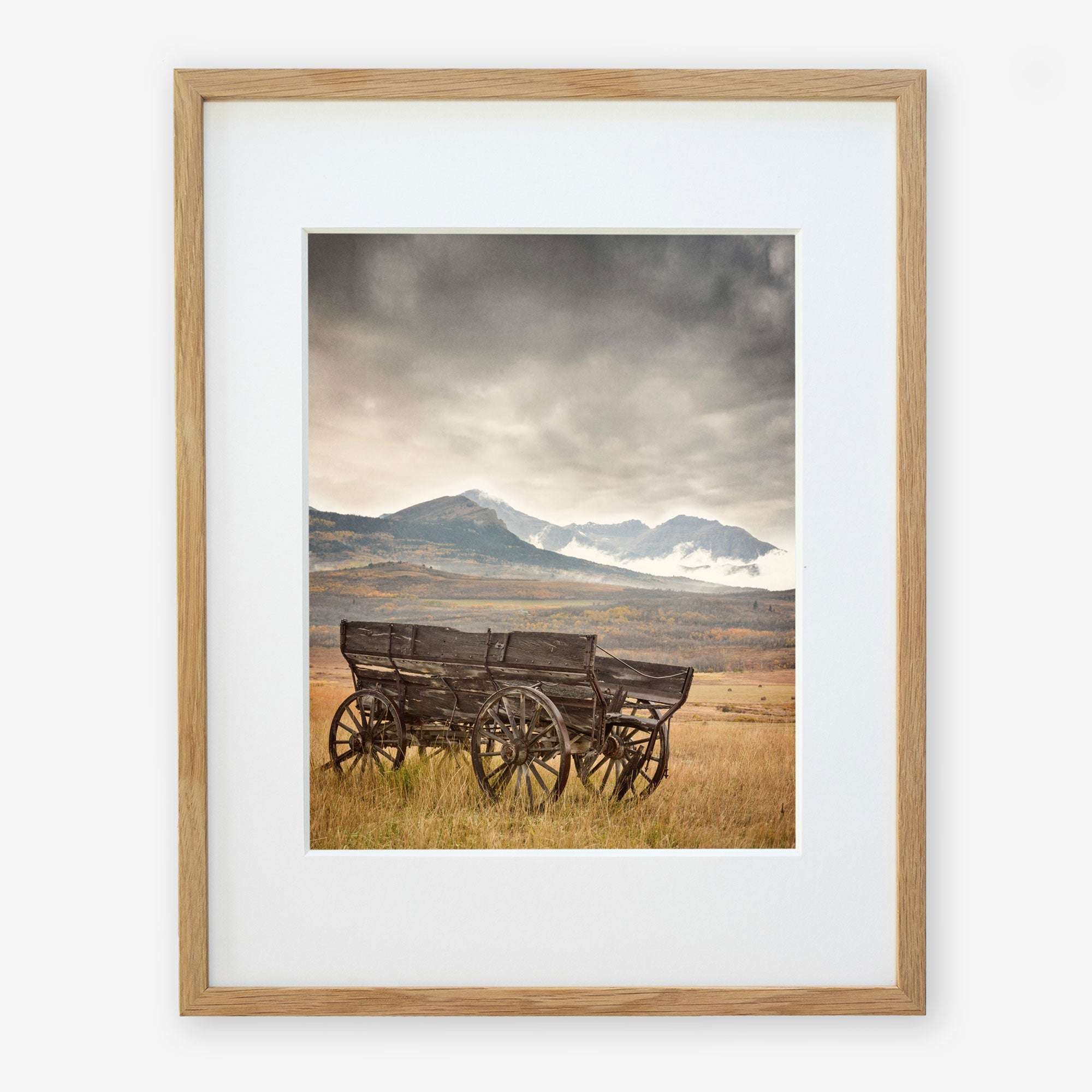 The "Old Wagon" by Offley Green is a Rustic Farmhouse Wall Art featuring a high-quality photograph of two wooden wagons, with one slightly weathered, on grassy plains under cloudy skies. Snow-capped mountains in the background add rustic charm, and it's elegantly framed with light wood and white matting.
