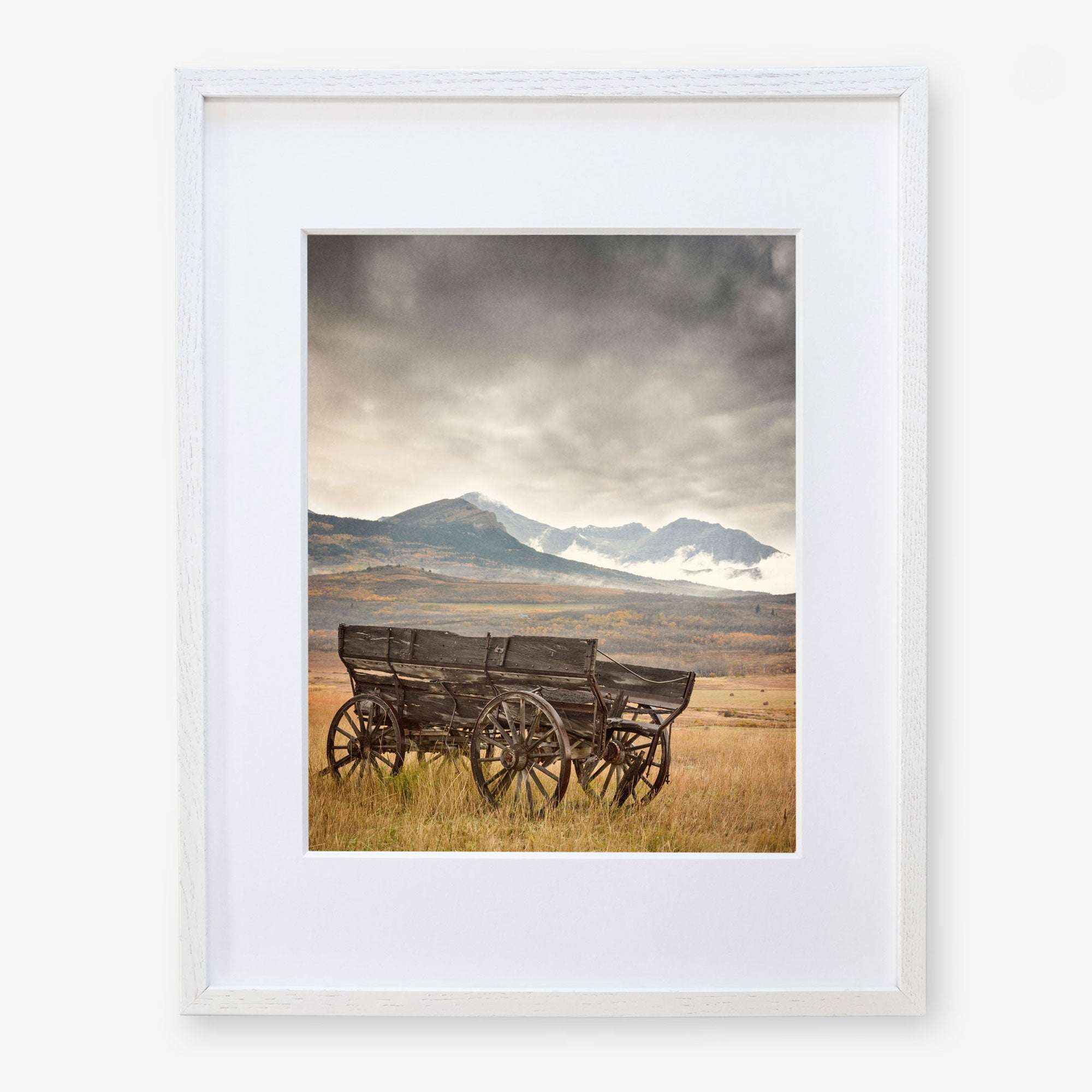 The Offley Green Rustic Farmhouse Wall Art, 'Old Wagon', features framed photography of vintage wooden wagons beneath a dramatic sky with snow-capped mountains, printed on premium photographic paper for a serene and rustic charm.