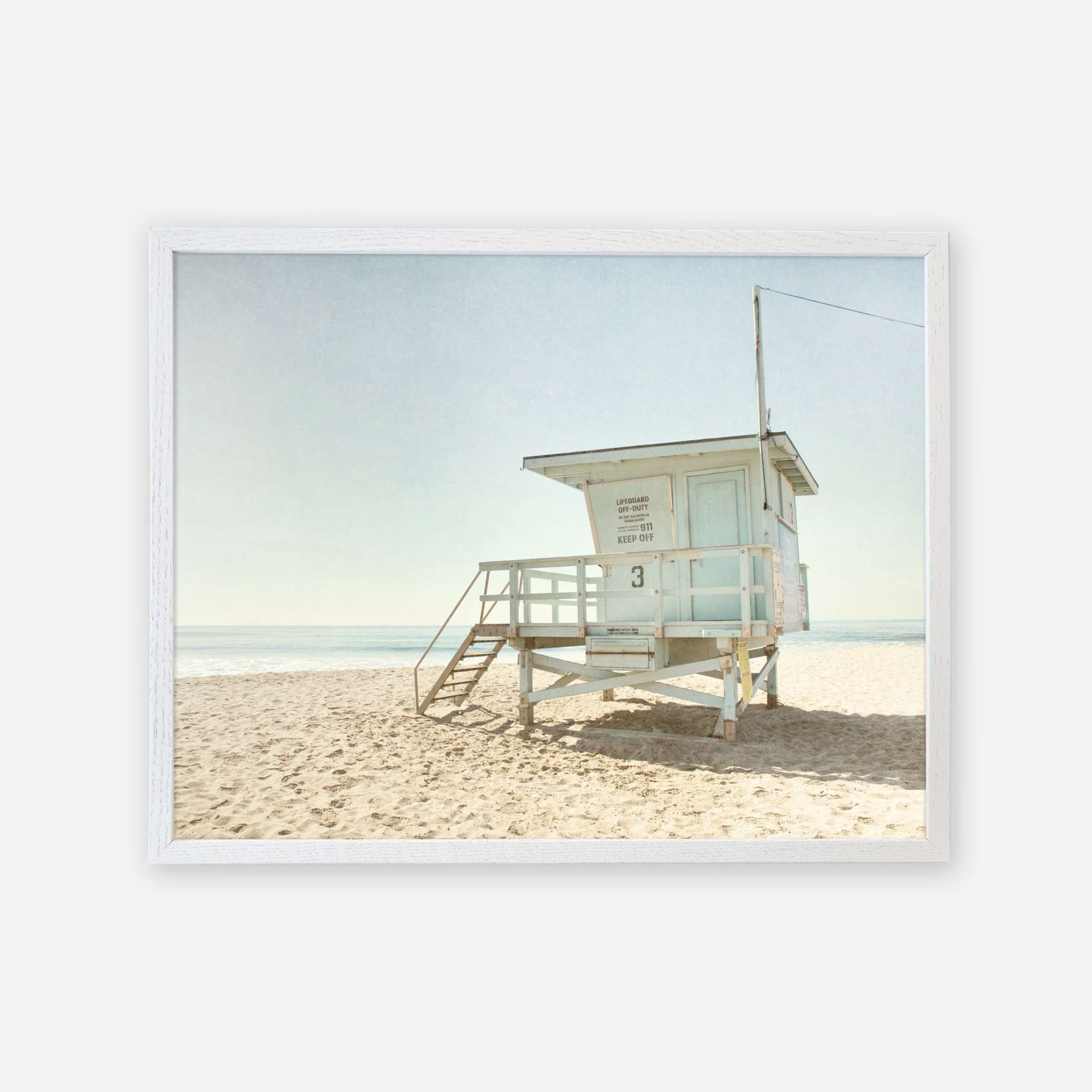 A framed picture of California Summer Beach Art, &#39;Malibu Lifeguard Tower&#39; by Offley Green on a serene beach under a clear sky. The sandy beach is empty, emphasizing the calmness of the setting.