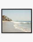 A framed wall art of a Malibu Beach House Print, 'Ocean View' by Offley Green, showing beach houses perched along the edge of a sandy beach with gentle waves lapping the shore under a clear sky.