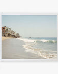 A framed wall art depicting a serene Malibu coastline with gentle waves, showcasing a row of houses built along the sandy shore under a clear sky. Offley Green's Malibu Beach House Print, 'Ocean View'.