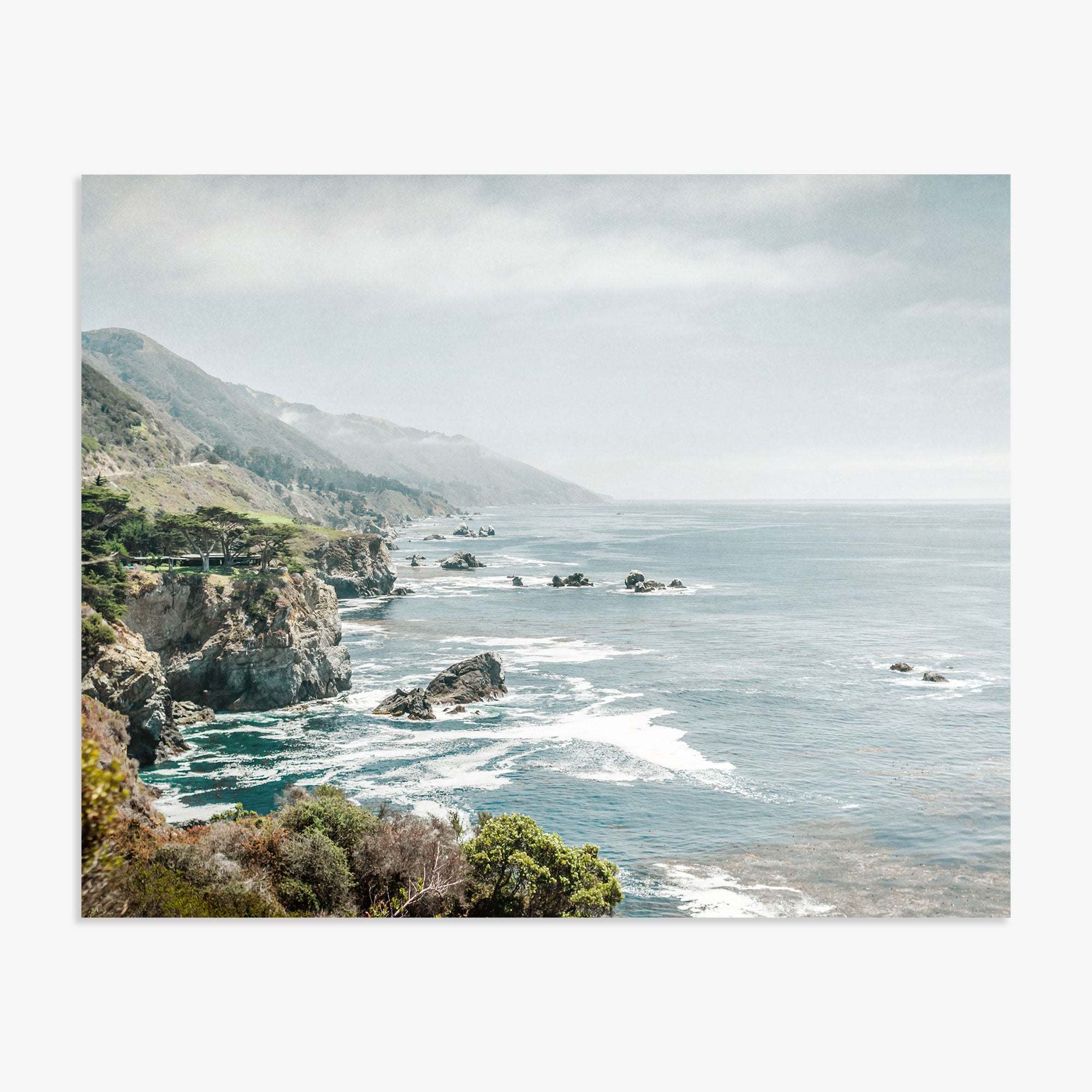 A scenic view of a misty coastline along California Highway 1 with rugged cliffs and waves crashing against the shore, under a soft cloudy sky. The landscape includes lush greenery and distant rocky outc featuring the Offley Green Big Sur Landscape Print, 'Rocky Rocks'.