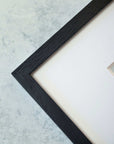 A close-up view of a corner of a black textured Offley Green picture frame against a light background, showcasing the frame's angle and a glimpse of an unframed Green Botanical Print, 'Strands and Spikes II' inside.