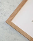 Close-up view of a wooden Offley Green picture frame corner on a textured gray background, showcasing the frame's natural wood grain and archival Malibu Beach House Print, 'Ocean View' paper inside the frame.