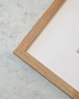 Close-up of a corner of a wooden picture frame on a marble surface, showing detail of the frame's texture and the clean, white archival photographic paper border inside it featuring Offley Green's Mint Green Botanical Print, 'Aloe Vera Spikes'.