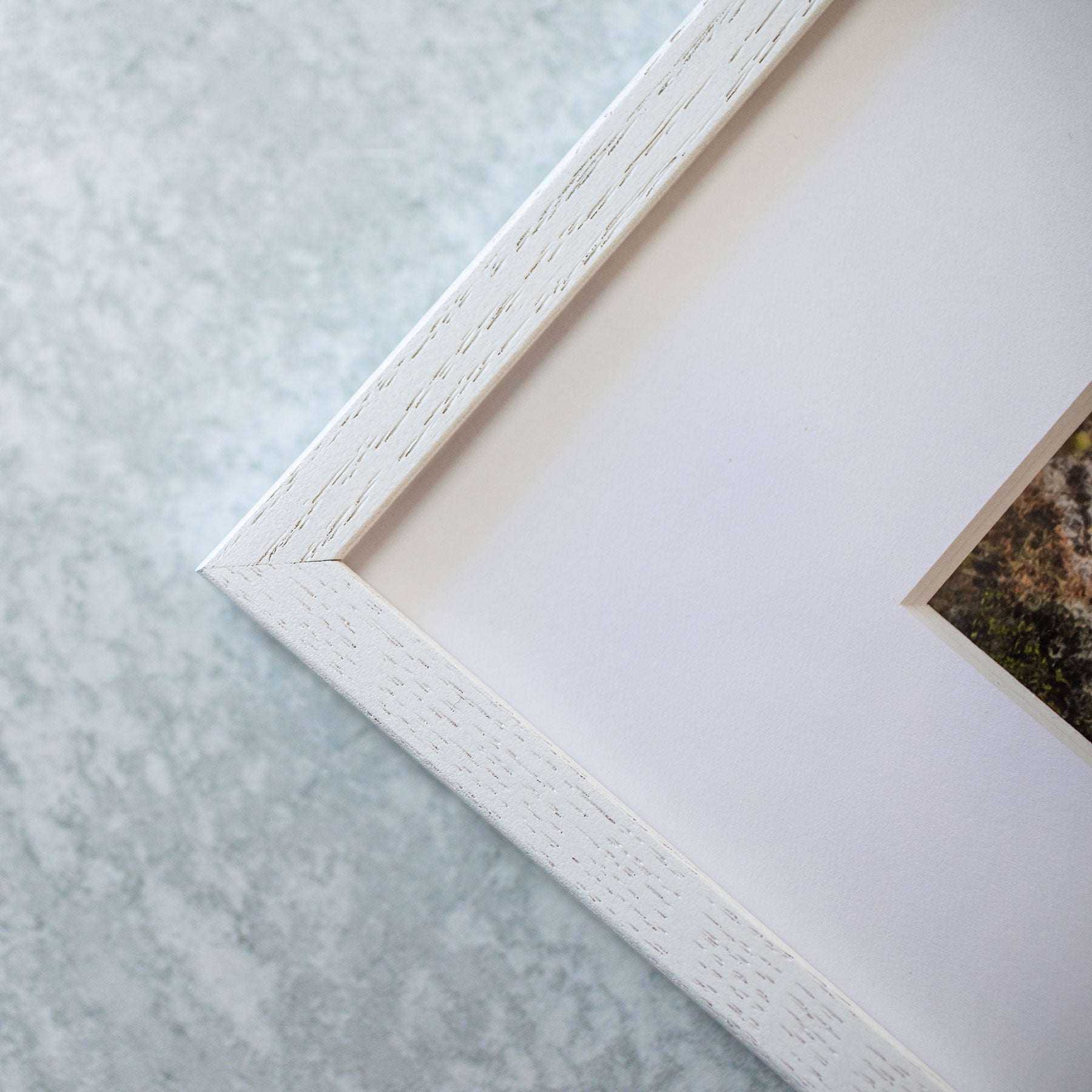 Close-up of the corner of Offley Green's Rustic Farmhouse Wall Art, 'Old Wagon,' against a textured gray background. The white wooden frame with visible grain adds a vintage touch to the partially visible, muted image it contains.