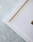 Close-up of the corner of Offley Green's Rustic Farmhouse Wall Art, 'Old Wagon,' against a textured gray background. The white wooden frame with visible grain adds a vintage touch to the partially visible, muted image it contains.