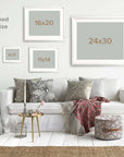 A cozy living room with a white sofa adorned with cushions, framed Offley Green Malibu Beach House Print, 'Ocean View' on archival photographic paper, a side table, lamp, and plants, presenting a comfortable and stylish interior.