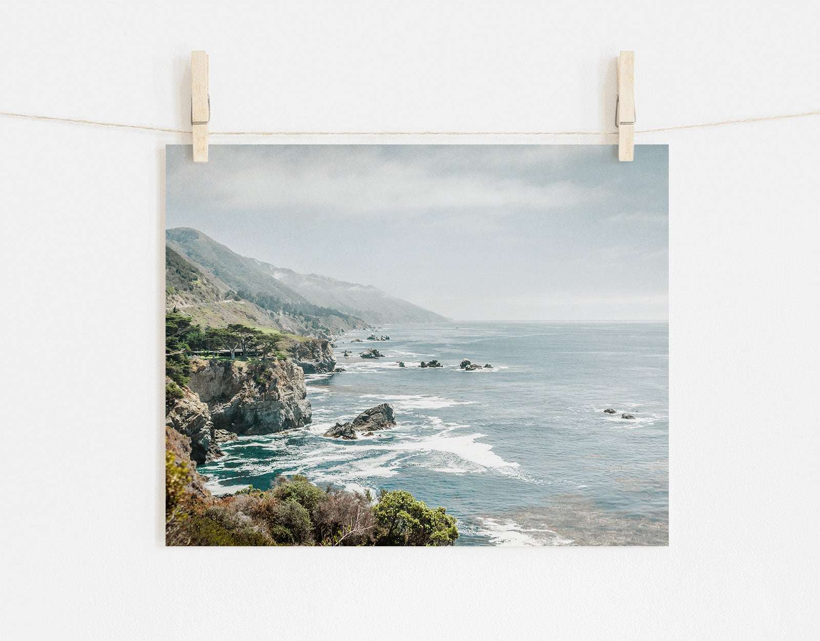 A photo clipped to a line, displaying a scenic view of Offley Green's 'Rocky Rocks' landscape print of Big Sur's rugged coastline with cliffs and ocean, wrapped in a gentle mist, under a pale sky.