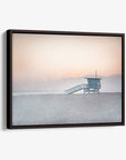A framed Pink Coastal Wall Art of a lifeguard station at Venice Santa Monica Beach, with soft pastel hues in the sky and serene water, mounted on a white wall by Offley Green.