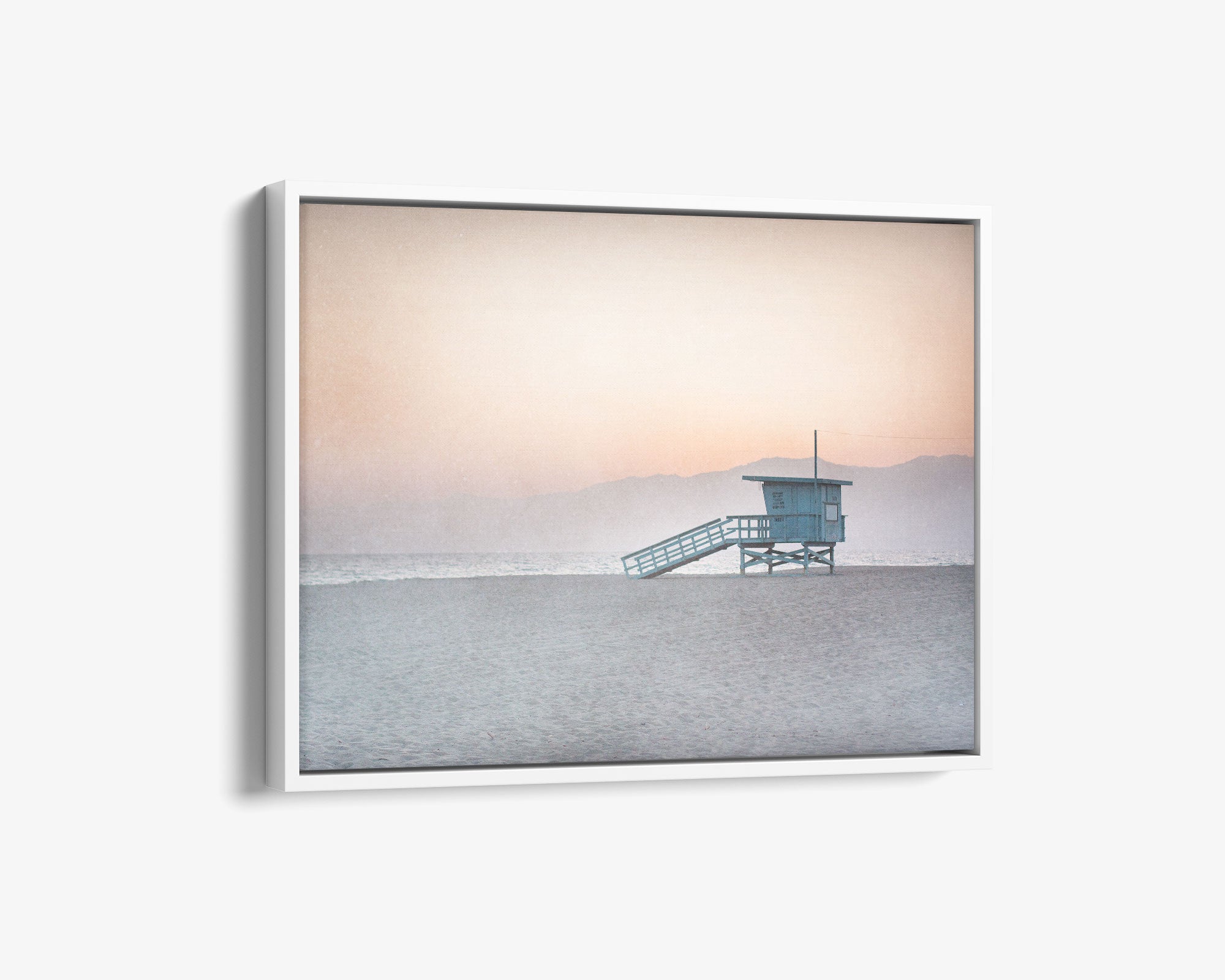A framed Pink Coastal Wall Art photograph of a 'Lifeguard Tower' on Venice Santa Monica Beach by Offley Green, with gentle hues of pink and blue in the sky and a soft glow reflecting off the ocean.