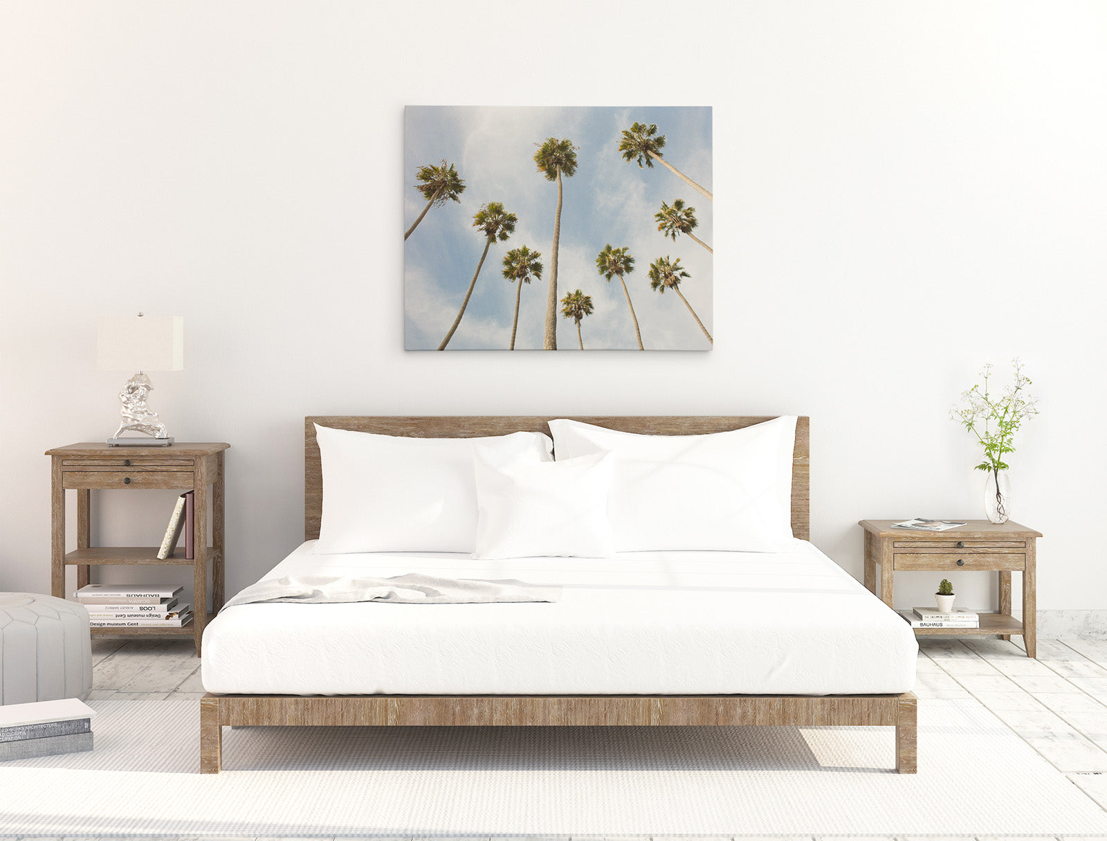 A minimalist bedroom features a wooden bed with white bedding, flanked by two wooden nightstands each holding a lamp. Centering the room is an Offley Green 30x40 California Canvas Print, depicting tall palm trees against a clear sky. Books and decor items adorn the space, creating a cozy atmosphere.
